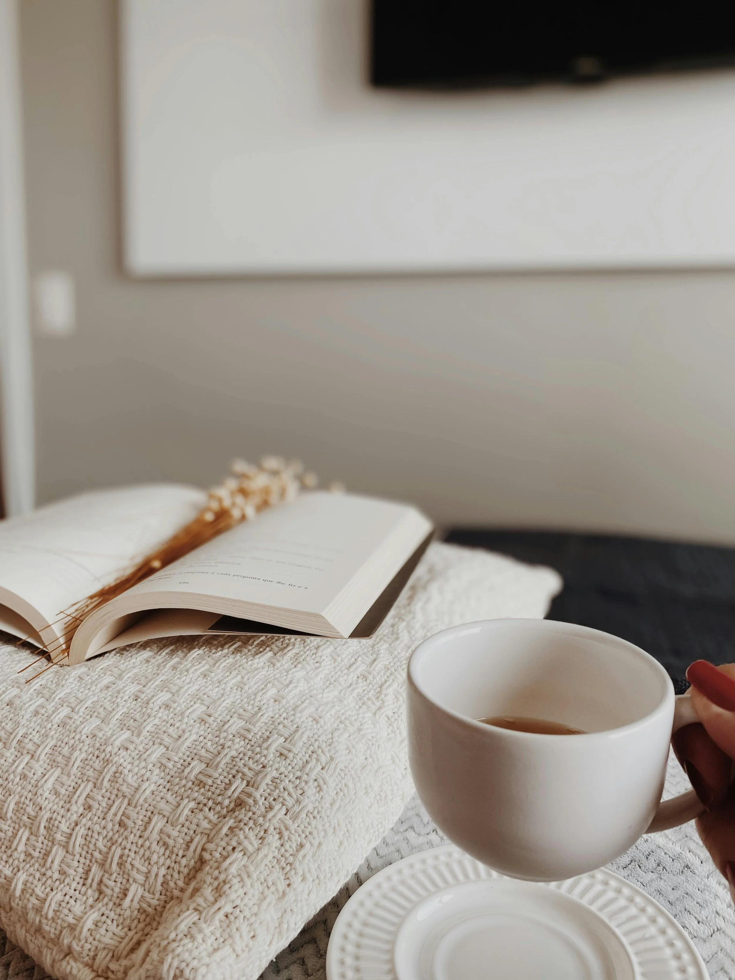 A cozy setting with an open book, a white ceramic mug of tea or coffee, and a white plate on a textured cream-colored tablecloth, with a blurred background of a TV and a white piece of furniture.
