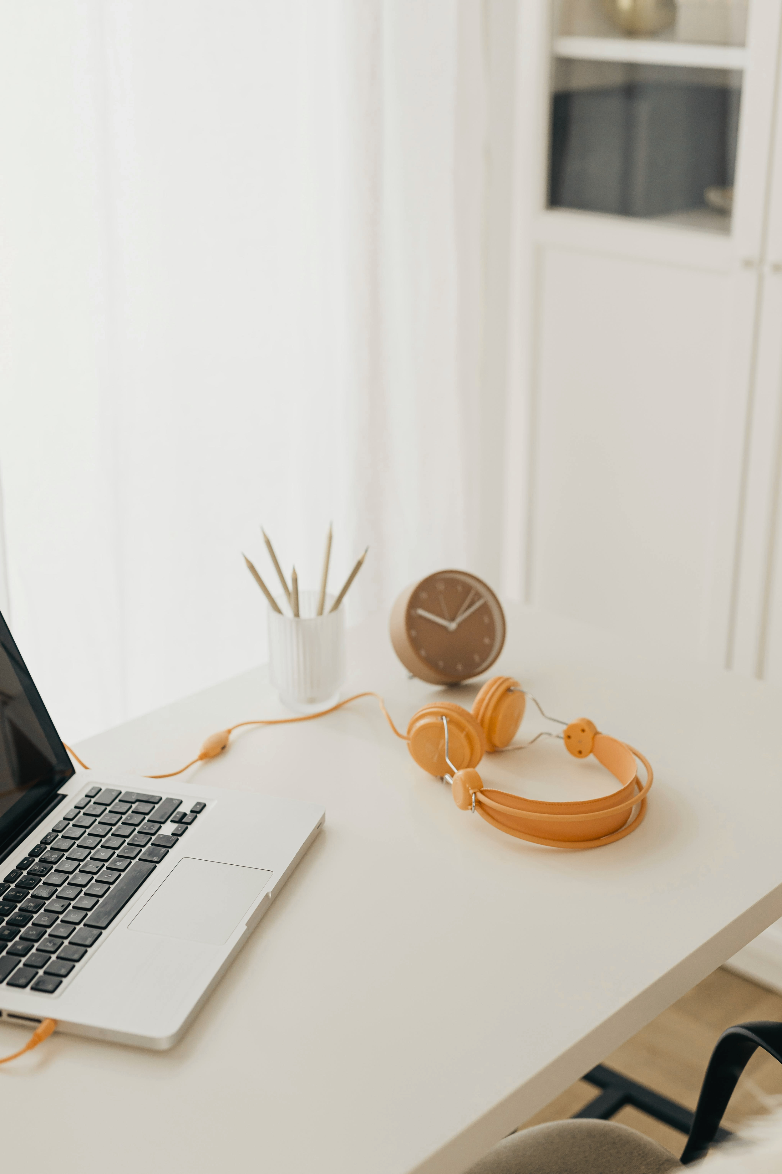 A white desk with an open laptop, orange over-ear headphones, a white vase with pencils, a brown round clock, and a beige office chair in a bright room with white walls and a solved cabinet in the background.