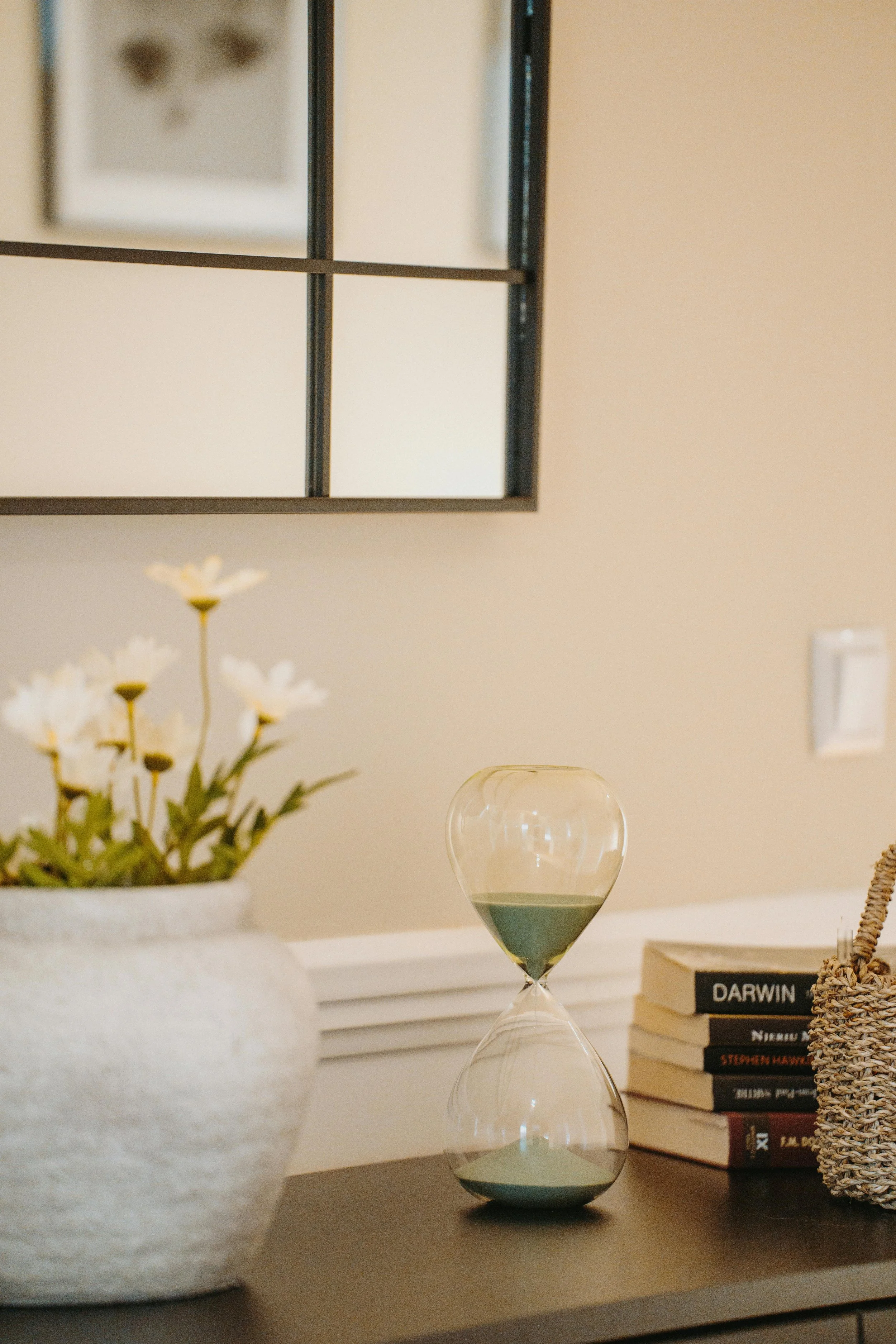 A decorative table setup with a flower vase, a stack of books, and a modern hourglass on a white surface, with a framed picture and cream-colored wall in the background.