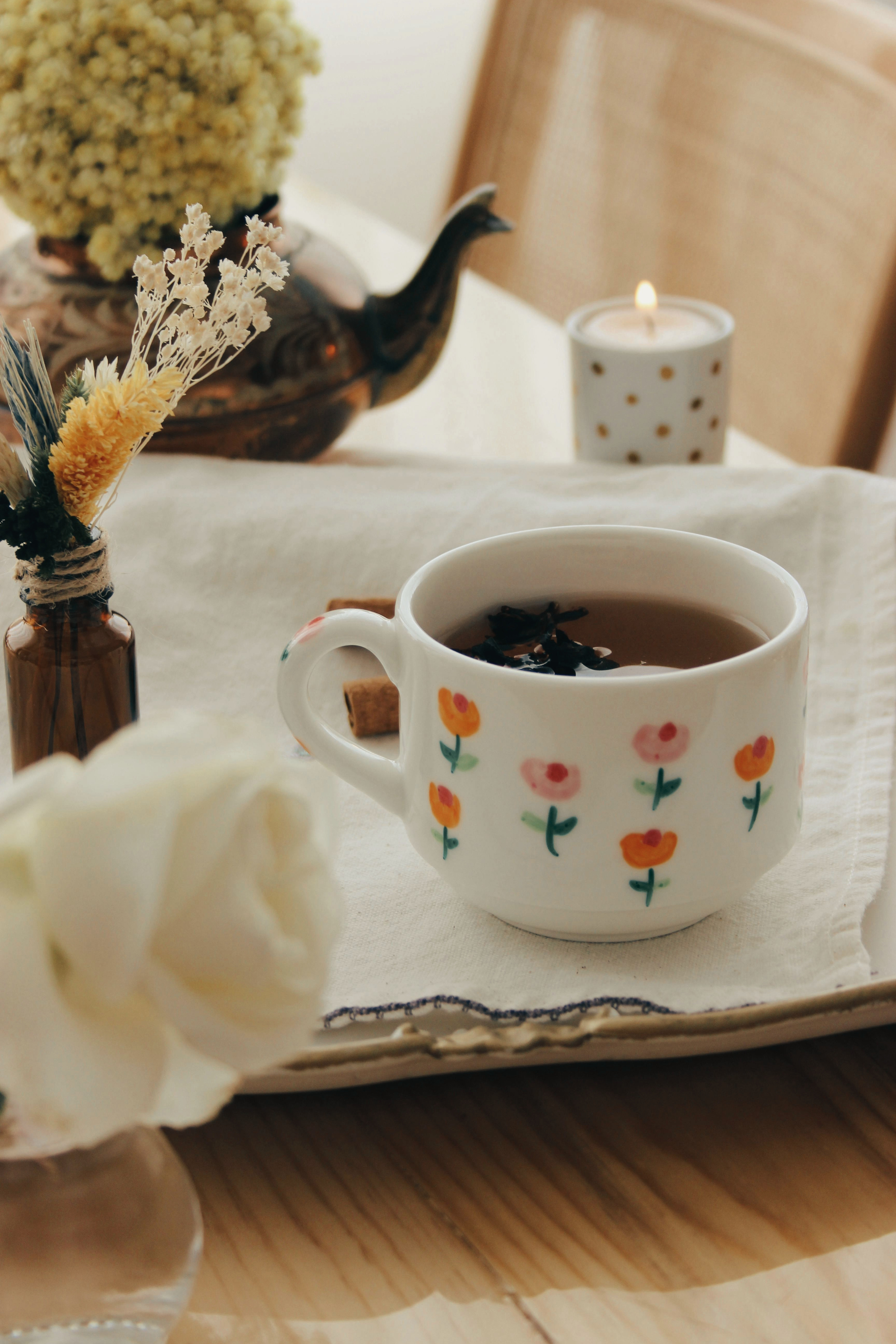 A decorated table with a cup of tea, a scented candle, a small vase with dried flowers, and a decorative bowl with a plant or flowers in it.