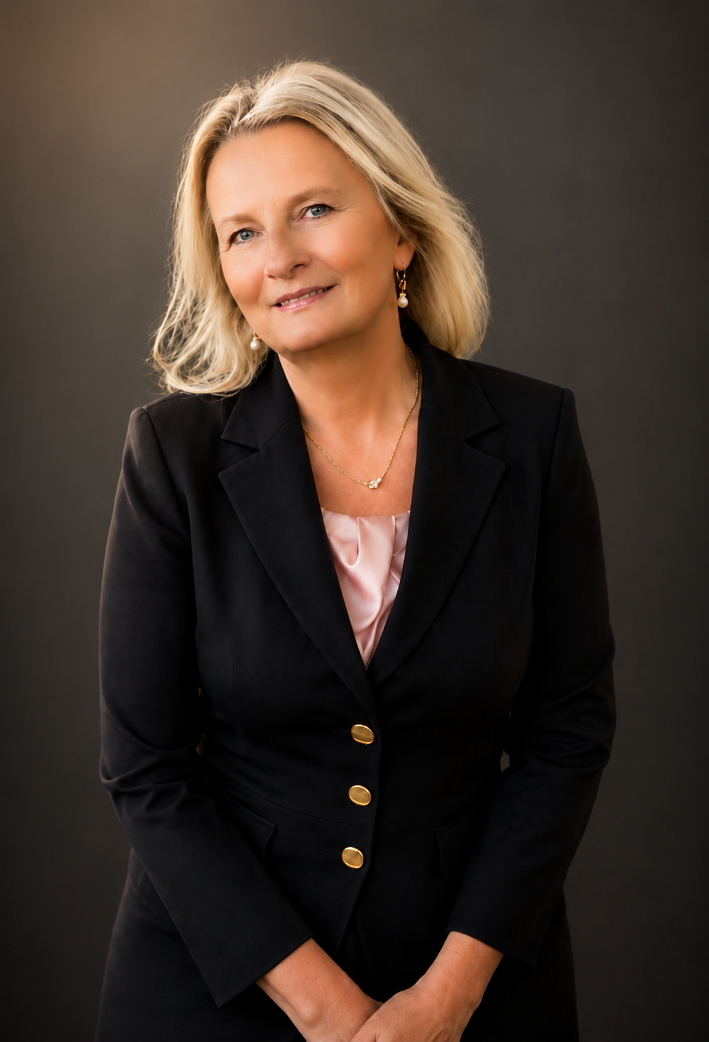 A portrait of a woman with blonde hair, wearing a black blazer with gold buttons and a pink satin blouse, accessorized with pearl earrings and a gold necklace, standing against a dark grey background.