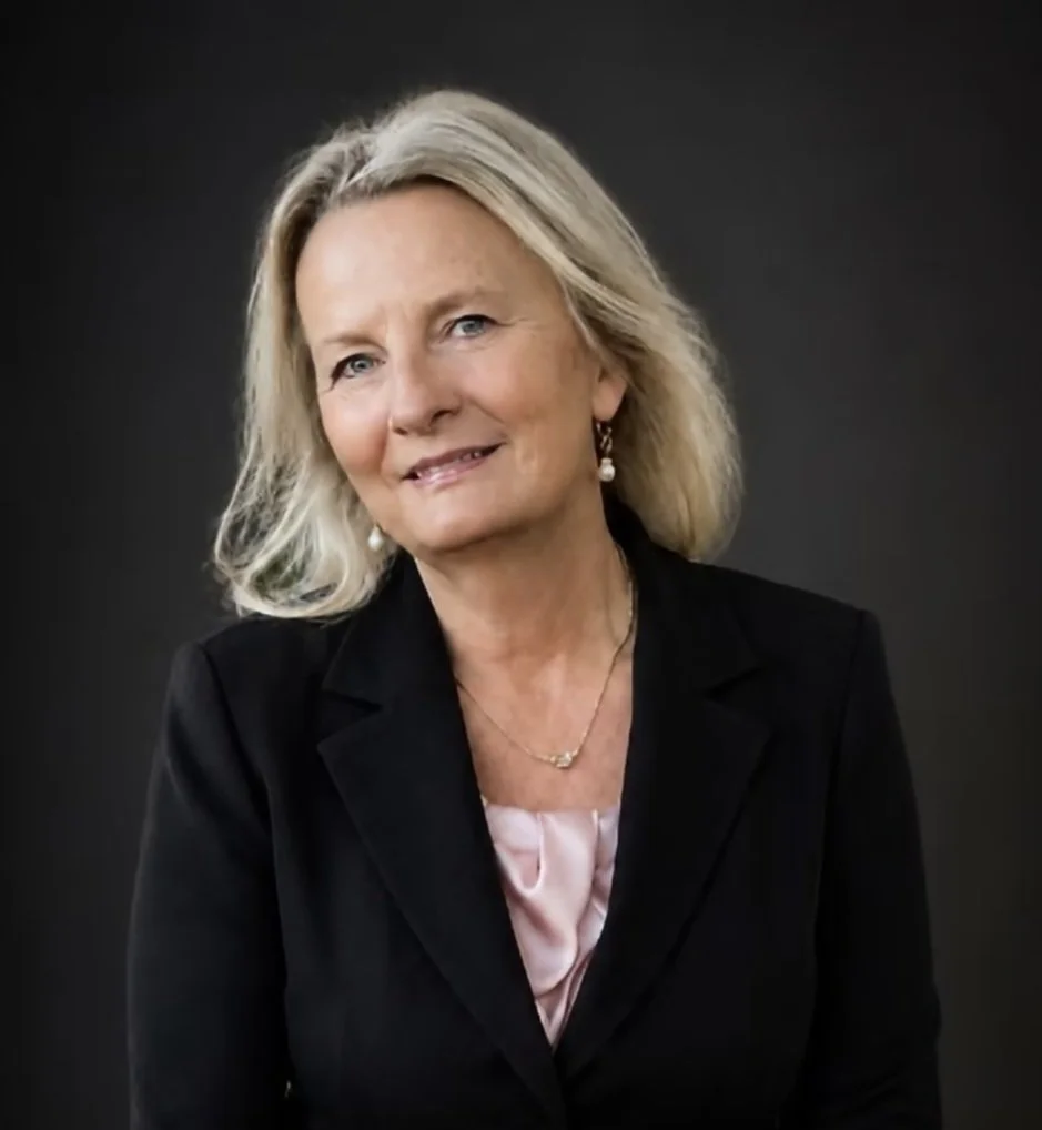 A portrait of a middle-aged woman with blonde hair wearing a black blazer, pink blouse, pearl earrings, and a gold necklace, against a dark background.