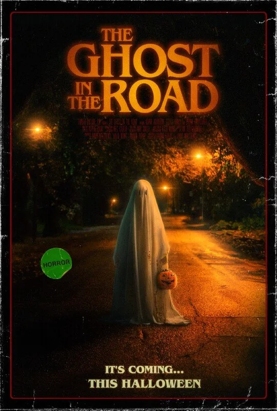 Movie poster for 'The Ghost in the Road' showing a ghostly figure covered with a white sheet holding a pumpkin-shaped bucket, standing on a dark, deserted street at night with streetlights, promoting a Halloween horror theme.