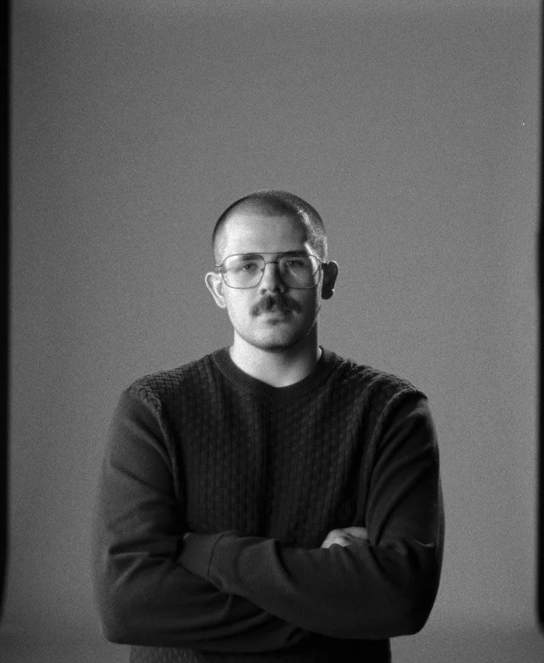 Black and white portrait of a man with glasses, a mustache, wearing a dark sweater, standing with arms crossed against a plain background.