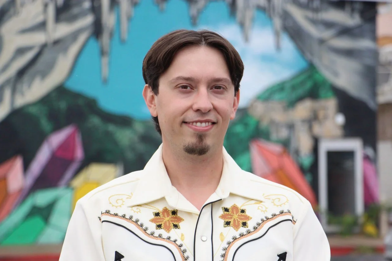 A man with dark hair, a goatee, and a mustache, smiling, wearing a white shirt with embroidered designs, standing in front of colorful murals.