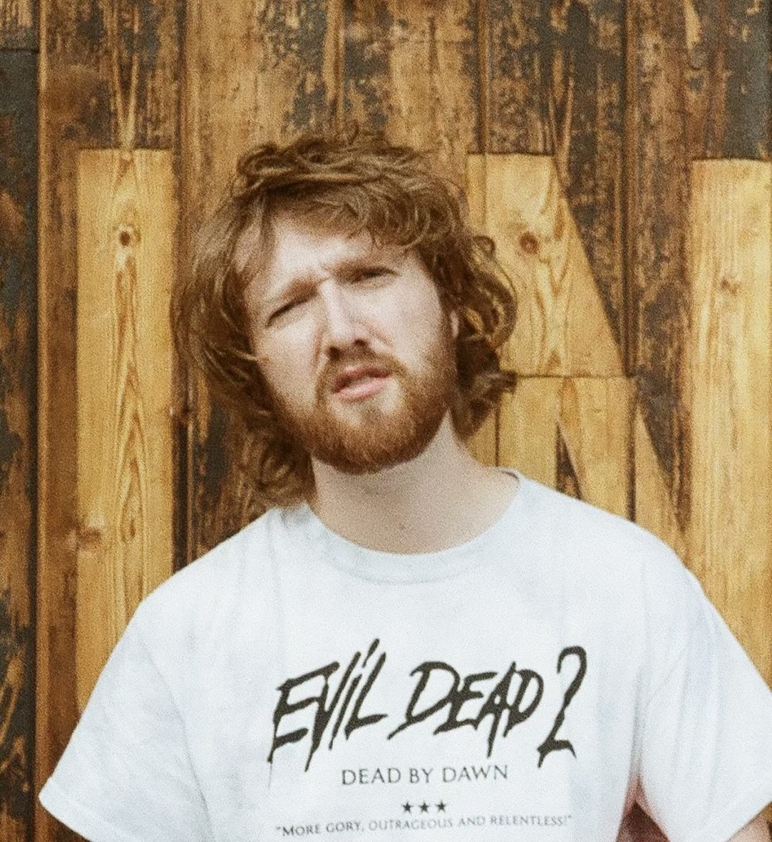 Man with red hair and beard making a funny face, wearing a white T-shirt with black text, standing in front of a wooden wall.