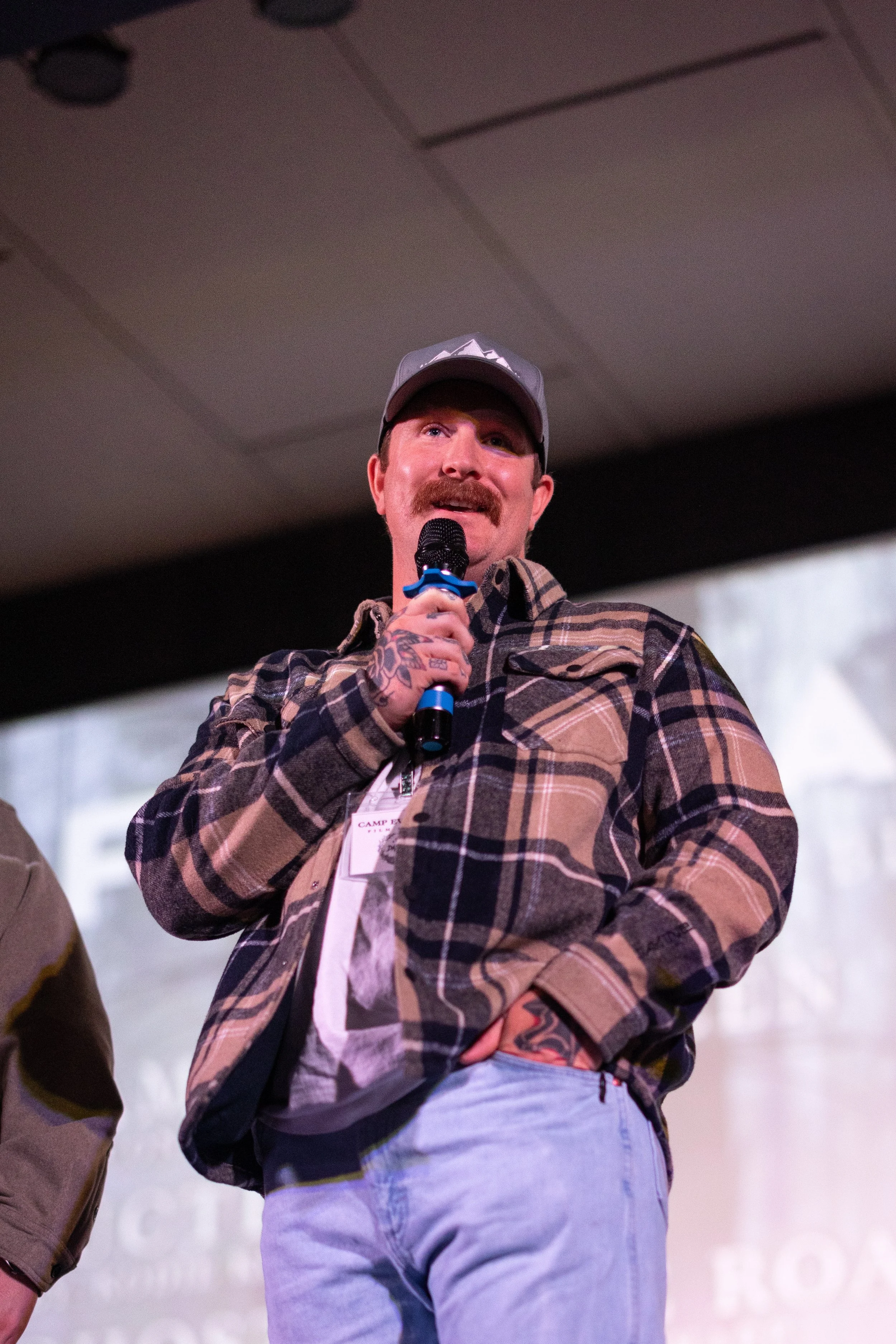A man with a goatee and mustache wearing a baseball cap, plaid shirt, and jeans holds a microphone and speaks on stage at an event.