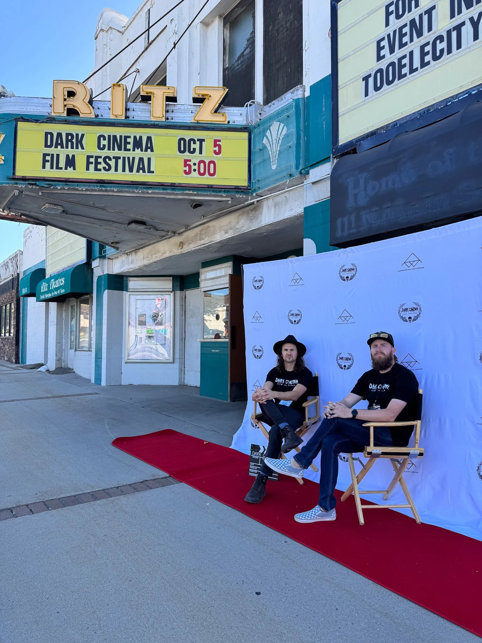 Two men sitting on director's chairs on a red carpet in front of a backdrop outside a theater. Signs indicate a Dark Cinema Film Festival event on October 5 at 5:00 PM.