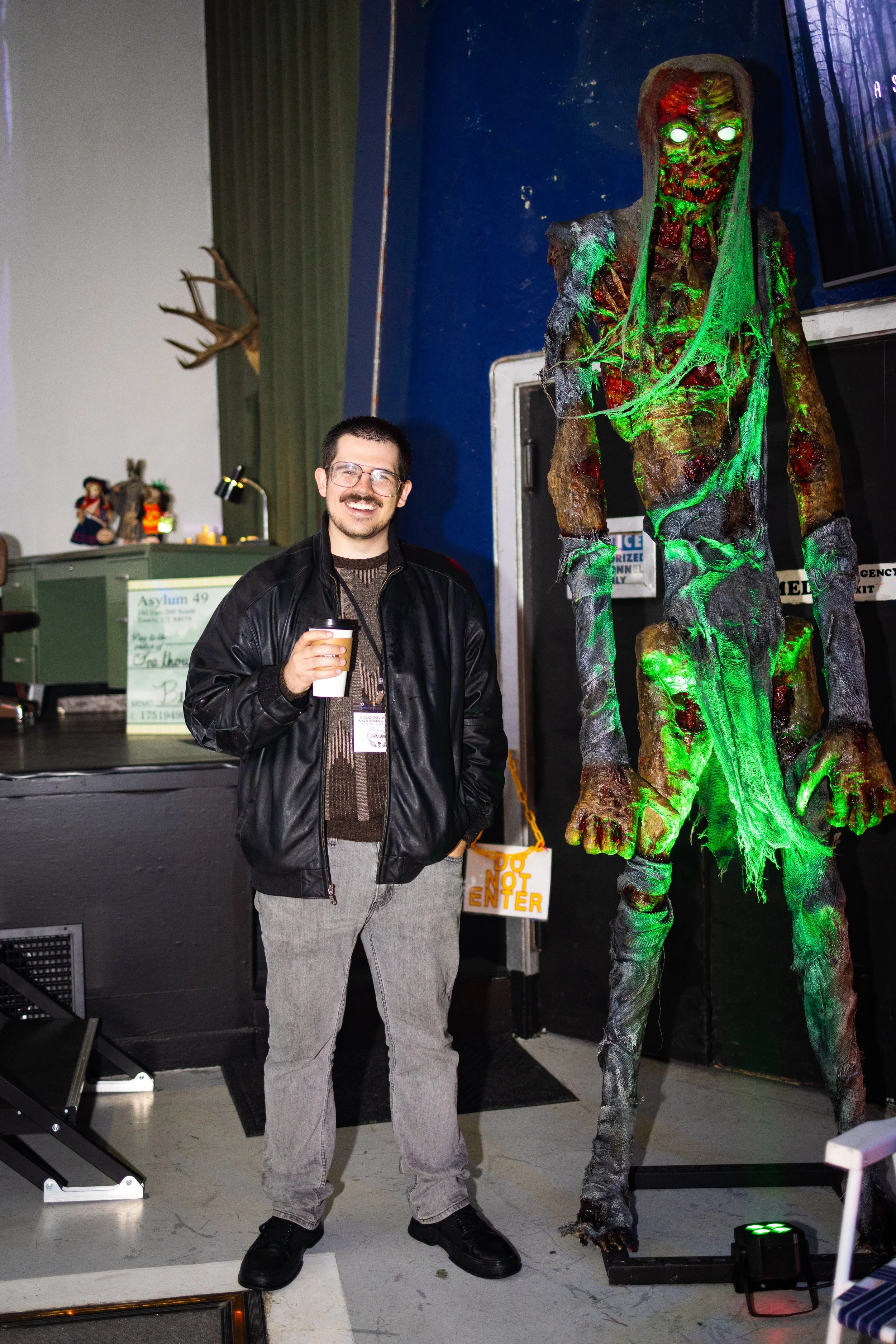 A man with glasses, a beard, and a brown sweater, holding a coffee cup, standing next to a large, creepy scarecrow-like figure with glowing green eyes and green lights on its body, in a dimly lit indoor space with dark blue walls and decorations.