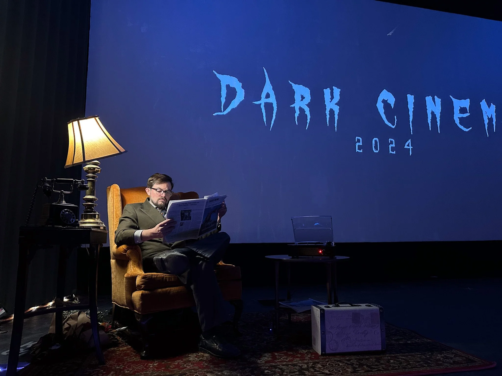 A man sitting in a vintage armchair, reading a newspaper in a dimly lit room. Next to him is a table with a retro telephone and a lamp. Behind him, a large screen displays "Dark Cinema 2024" in stylized text.