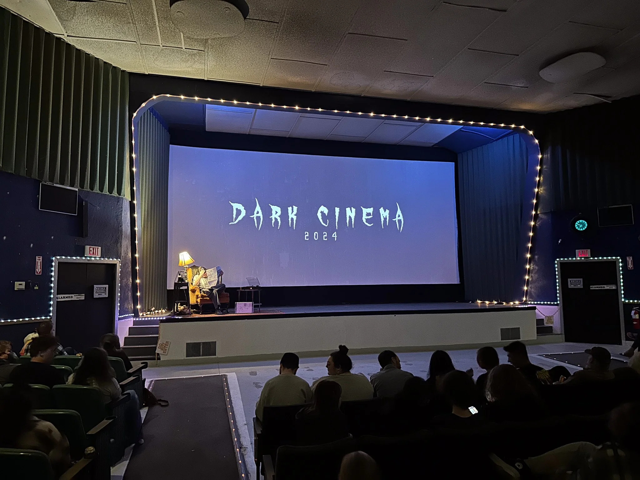 Audience seated in a dark auditorium watching a movie on a large screen with the title 'Dark Cinema 2024', decorated with fairy lights.