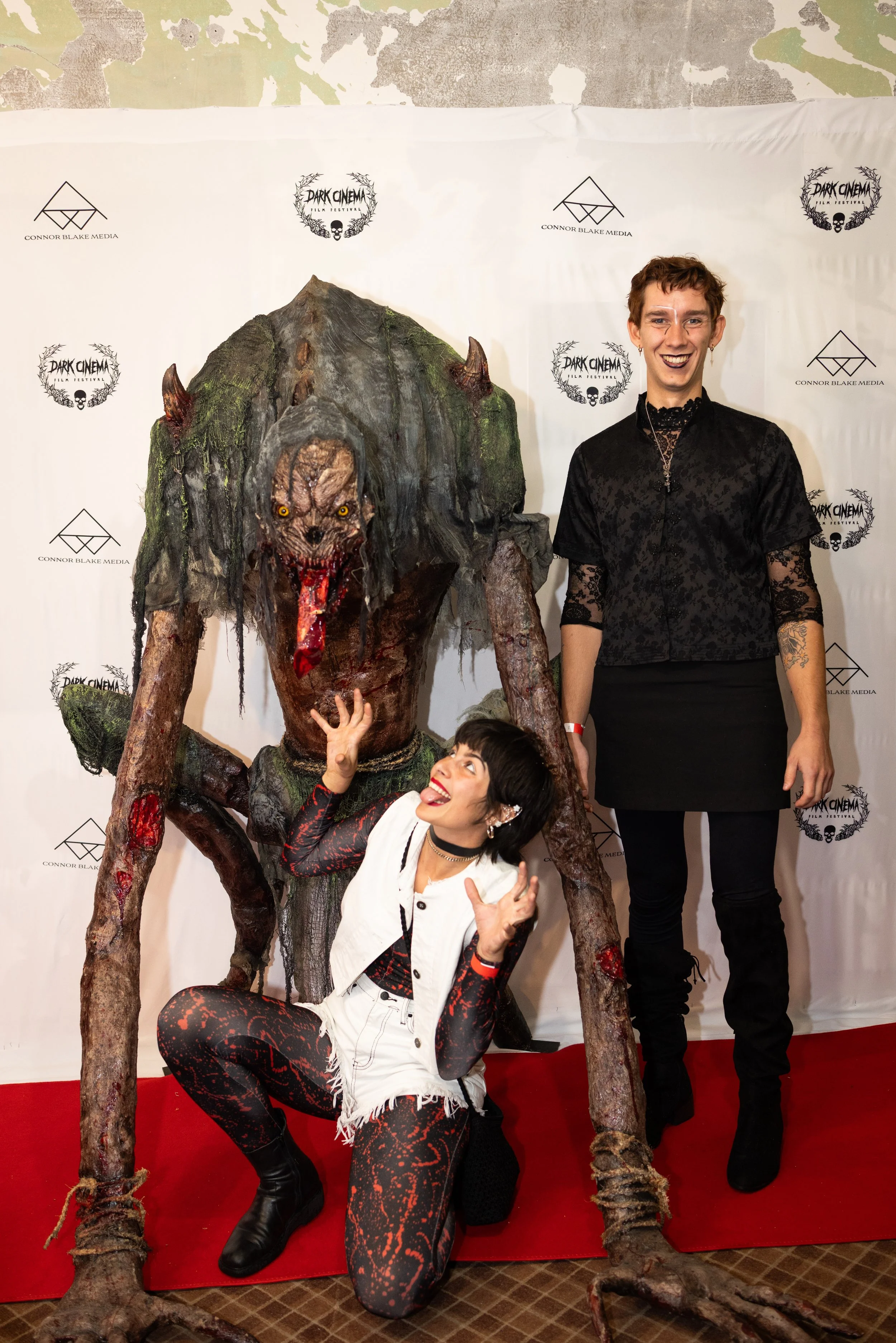 Three people posing with a large, menacing tree monster at a film event. The monster has a grotesque face with yellow eyes, a wide open mouth with sharp teeth and a tongue, long dreadlocked hair, and a mossy body. The background features a white bann