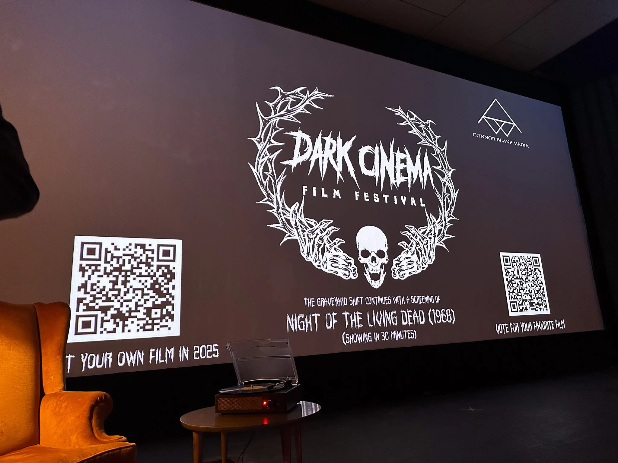A projection screen displaying a poster for the Dark Cinema Film Festival, featuring a skull with thorns, two skeletal hands, and QR codes on either side.