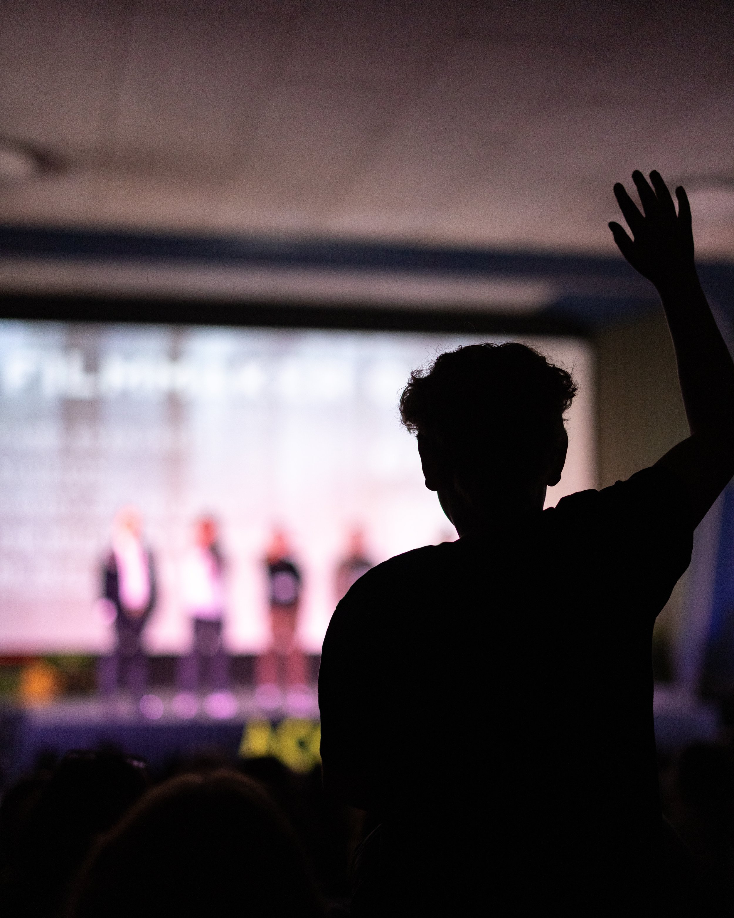A silhouette of a person raising their hand at a conference or event with a stage and four people on it in the background.