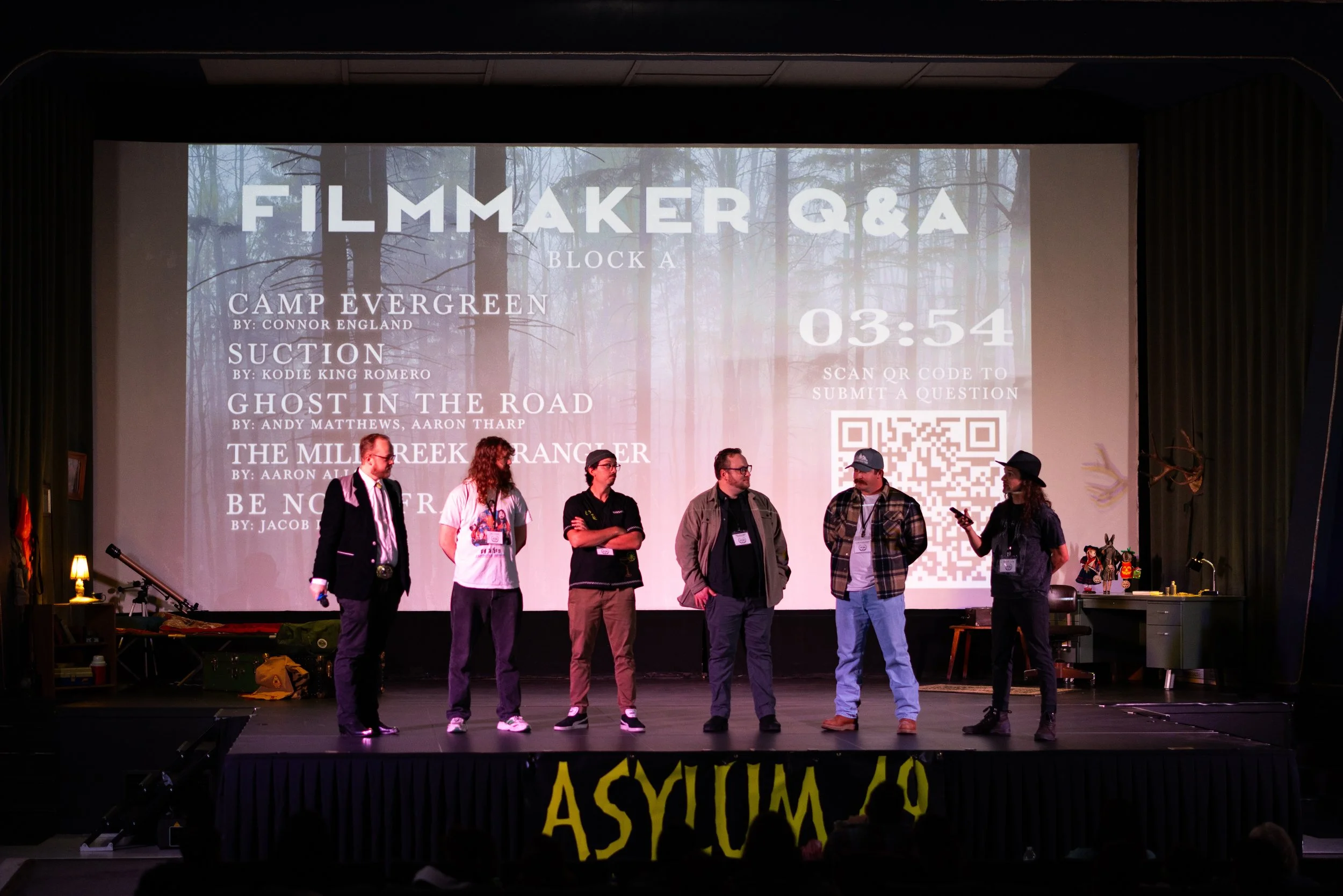 Panel of six people on stage during a Filmmaker Q&A event, with a large screen behind them displaying event details and a QR code.