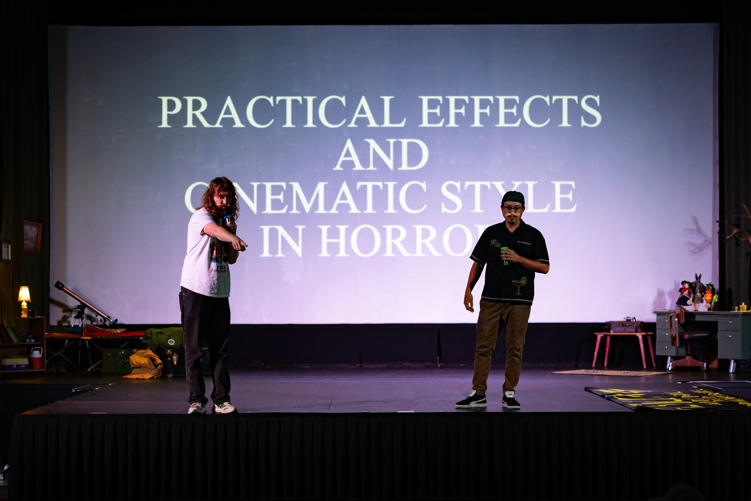 Two people standing on a stage, one holding a microphone, with a large screen behind them displaying the words 'Practical Effects and Cinematic Style in Horror'.