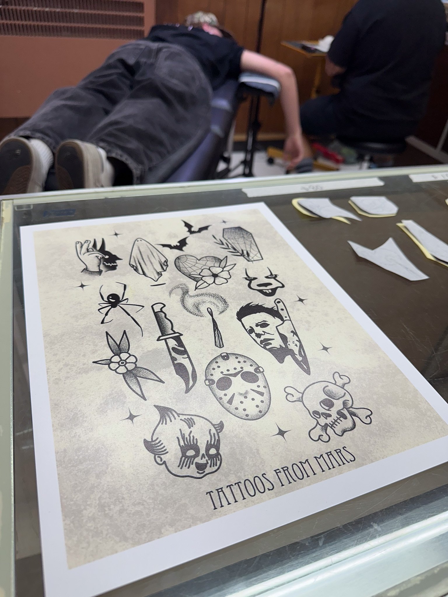 A sheet of temporary tattoo designs featuring images of bats, a ghost, a heart, a spider, a dagger, a face, a skull, a ghost face, a doll face, and a clock, with the text 'Tattoos from Mars' at the bottom. In the background, a person is lying face do
