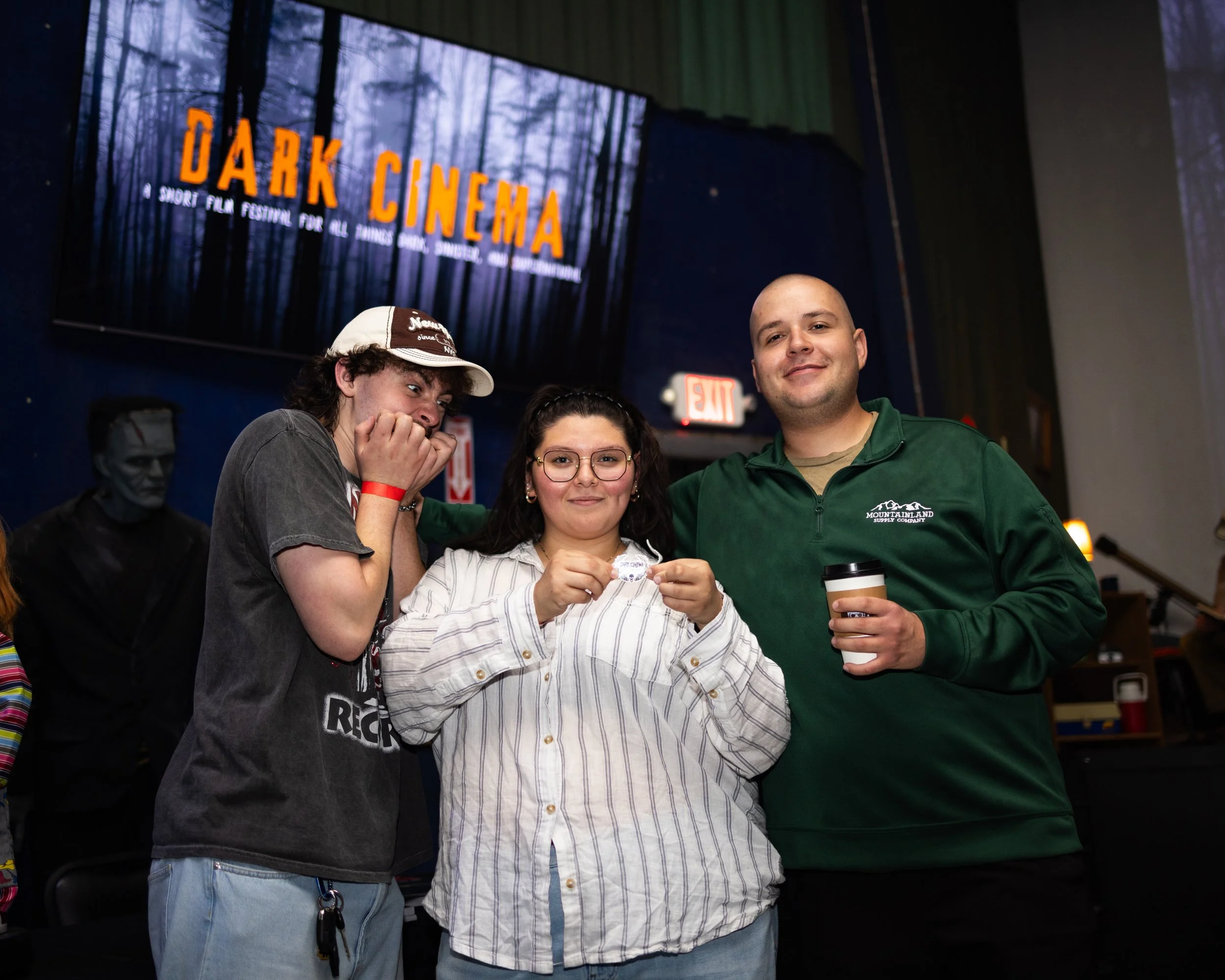 Three people standing together at a dark cinema event with a large screen in the background displaying 'Dark Cinema,' a forest scene, and a black forest backdrop. The person on the left is laughing and covering their mouth, the person in the middle i