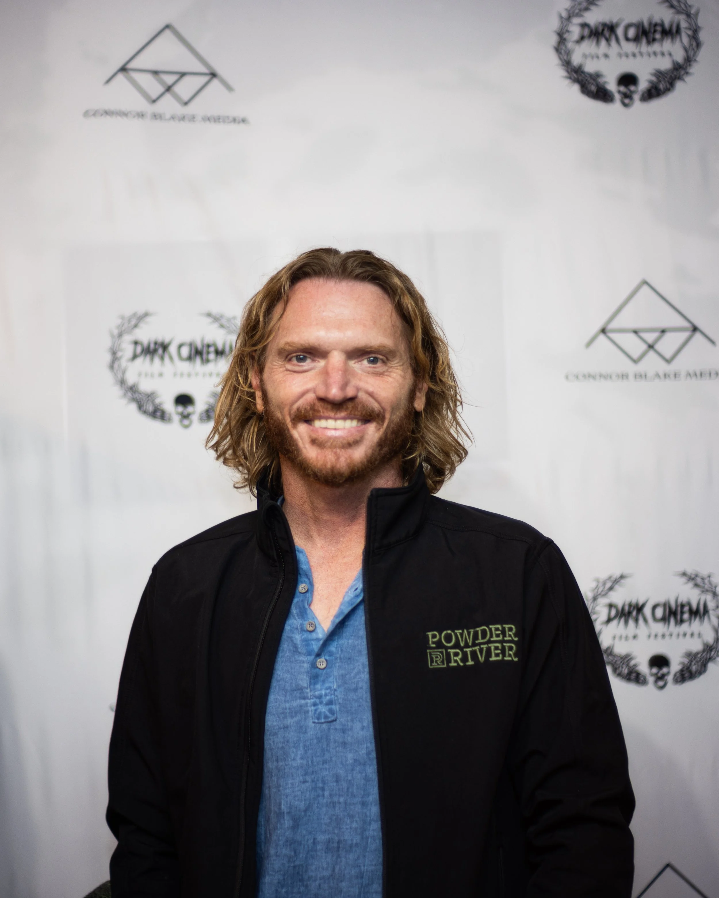 Smiling man with long wavy hair and a beard, wearing a black jacket with 'Powder River' logo, standing in front of a backdrop with logos and text, including 'Dark Omen' and 'Connor Blake Media'.