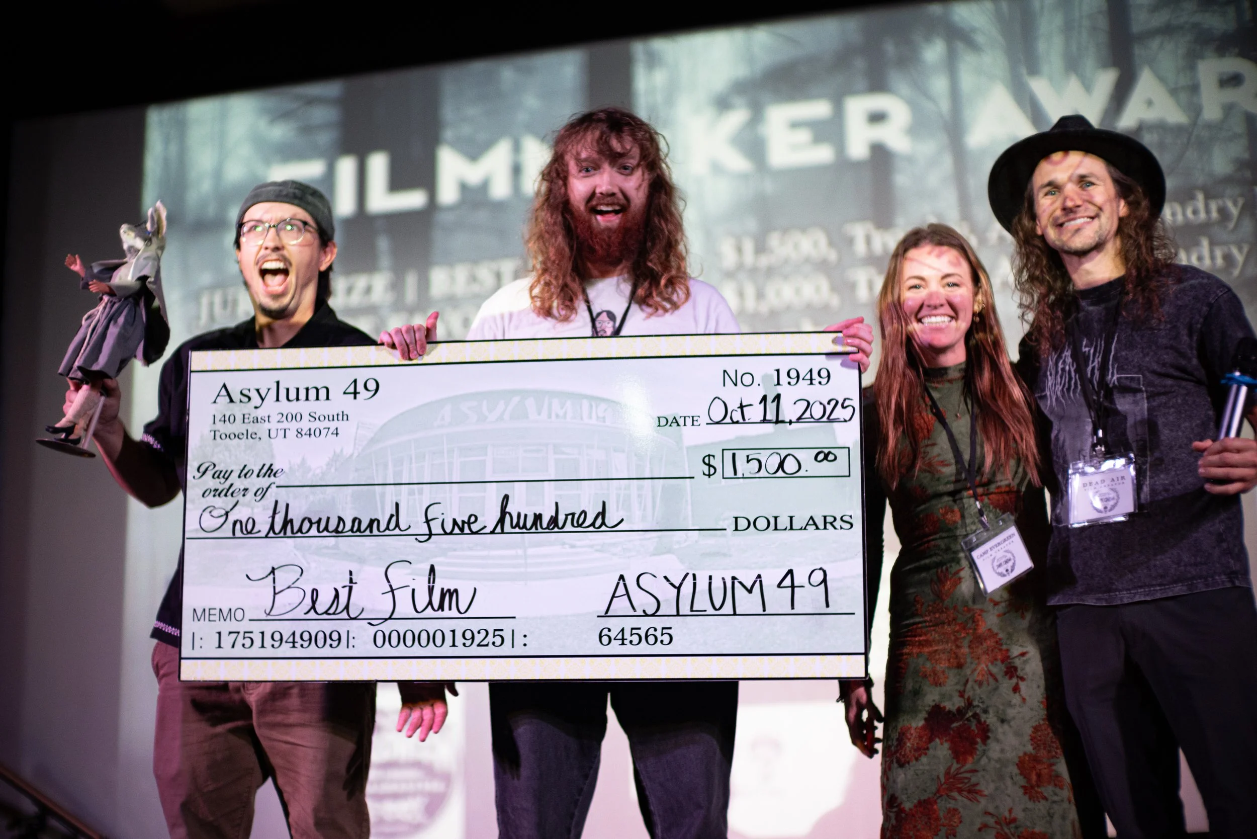 Group of four people on stage holding an oversized check for $1,500 made out to Best Film, with one person holding a puppet and another with a microphone, celebrating an award at the film festival.