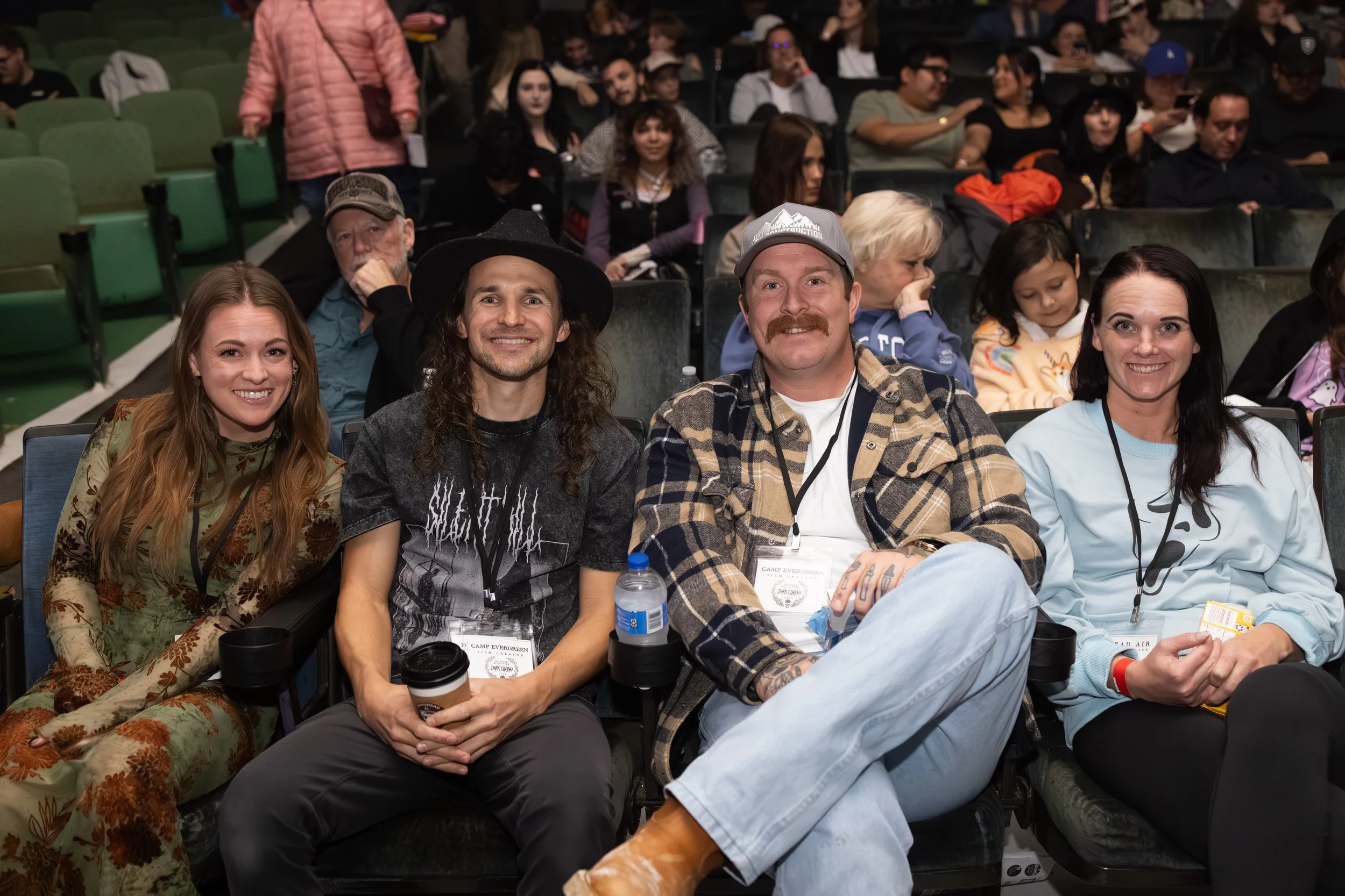 Four people sitting in an auditorium, smiling at the camera. The group includes two women and two men, with other audience members visible in the background.
