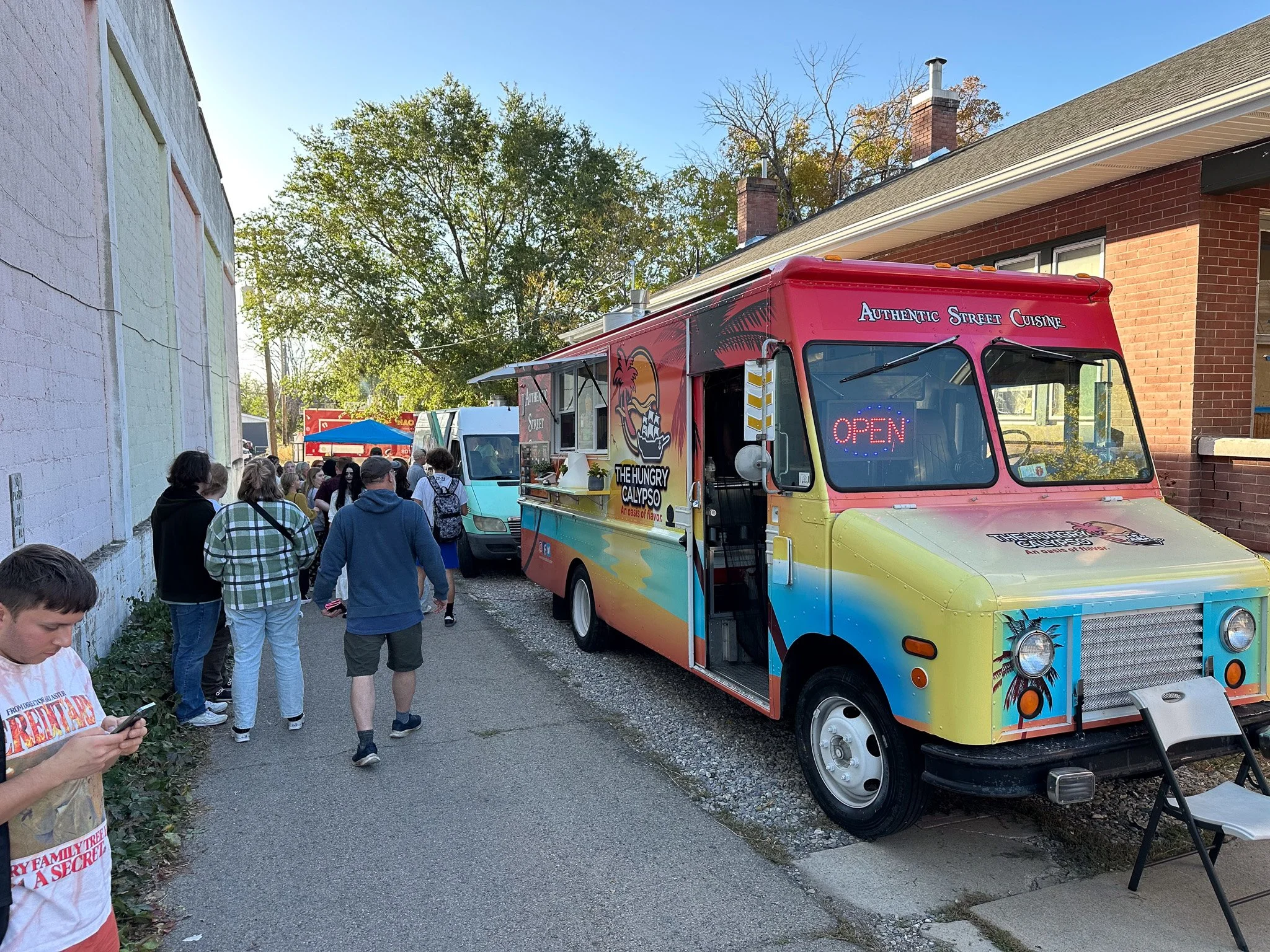 A colorful food truck with a sign that says 'Authentic Street Cuisine' and an illuminated 'Open' sign parked on a side street, with people lining up to order, and other trucks visible in the background on a sunny day.