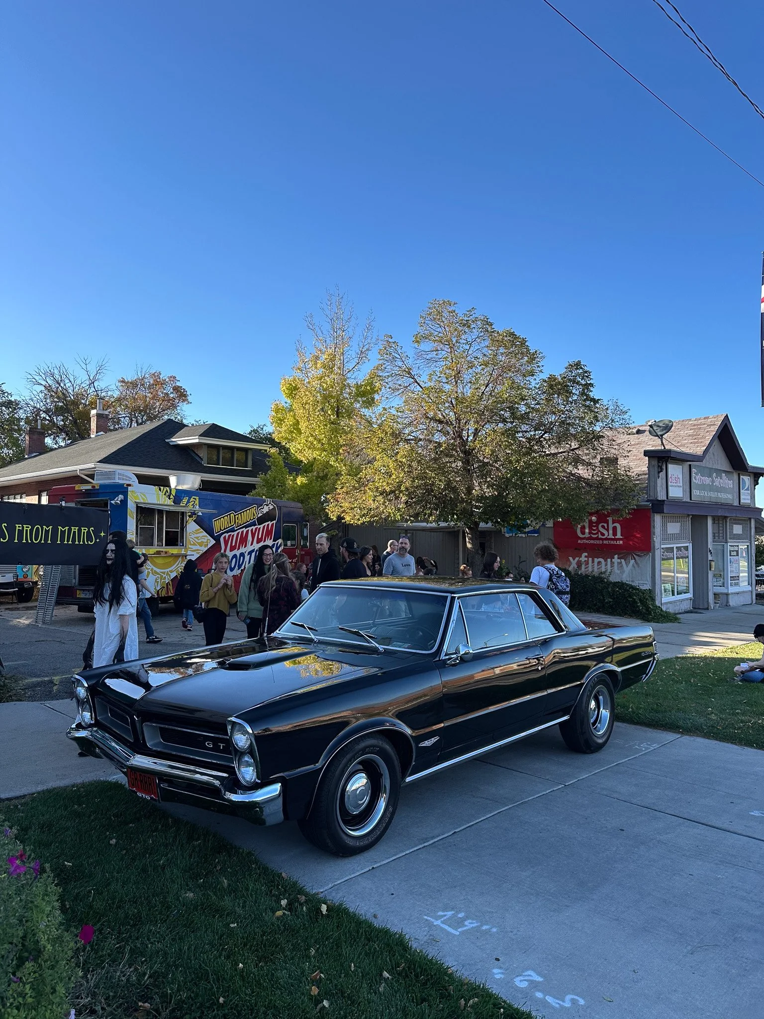 A vintage black car parked on the street with a crowd of people and a food truck in the background.