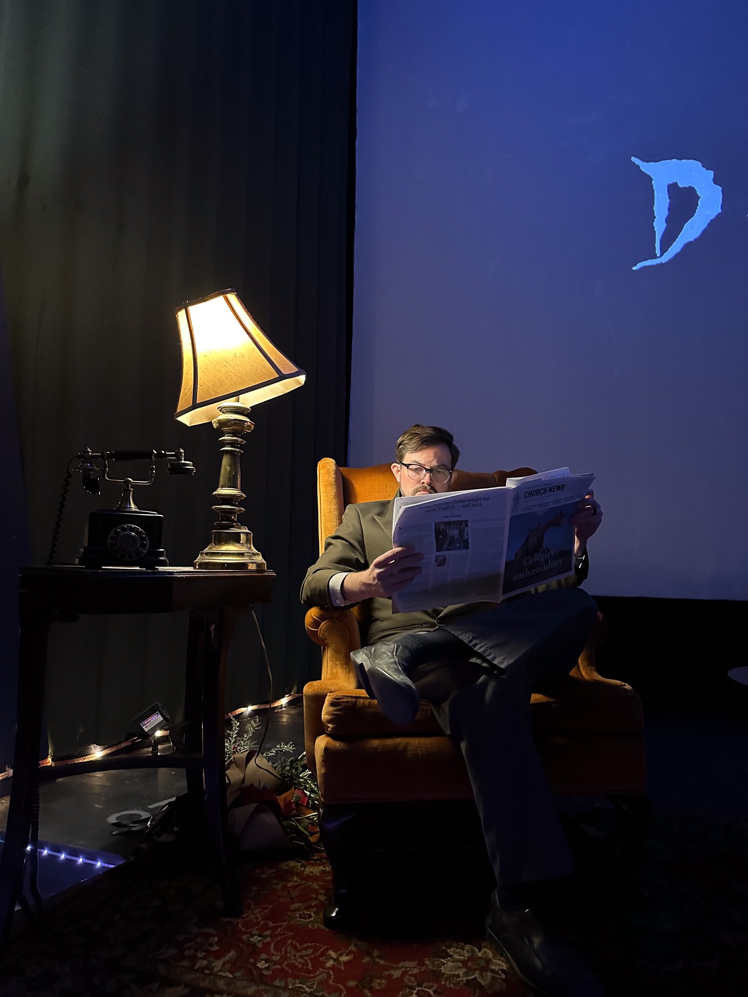 A man in glasses and a suit sitting on a brown armchair, reading a newspaper, beside a table with a lamp, a vintage rotary phone, and holiday decorations, in a dimly lit room with a large screen showing a blue background and a partial letter 'D'.