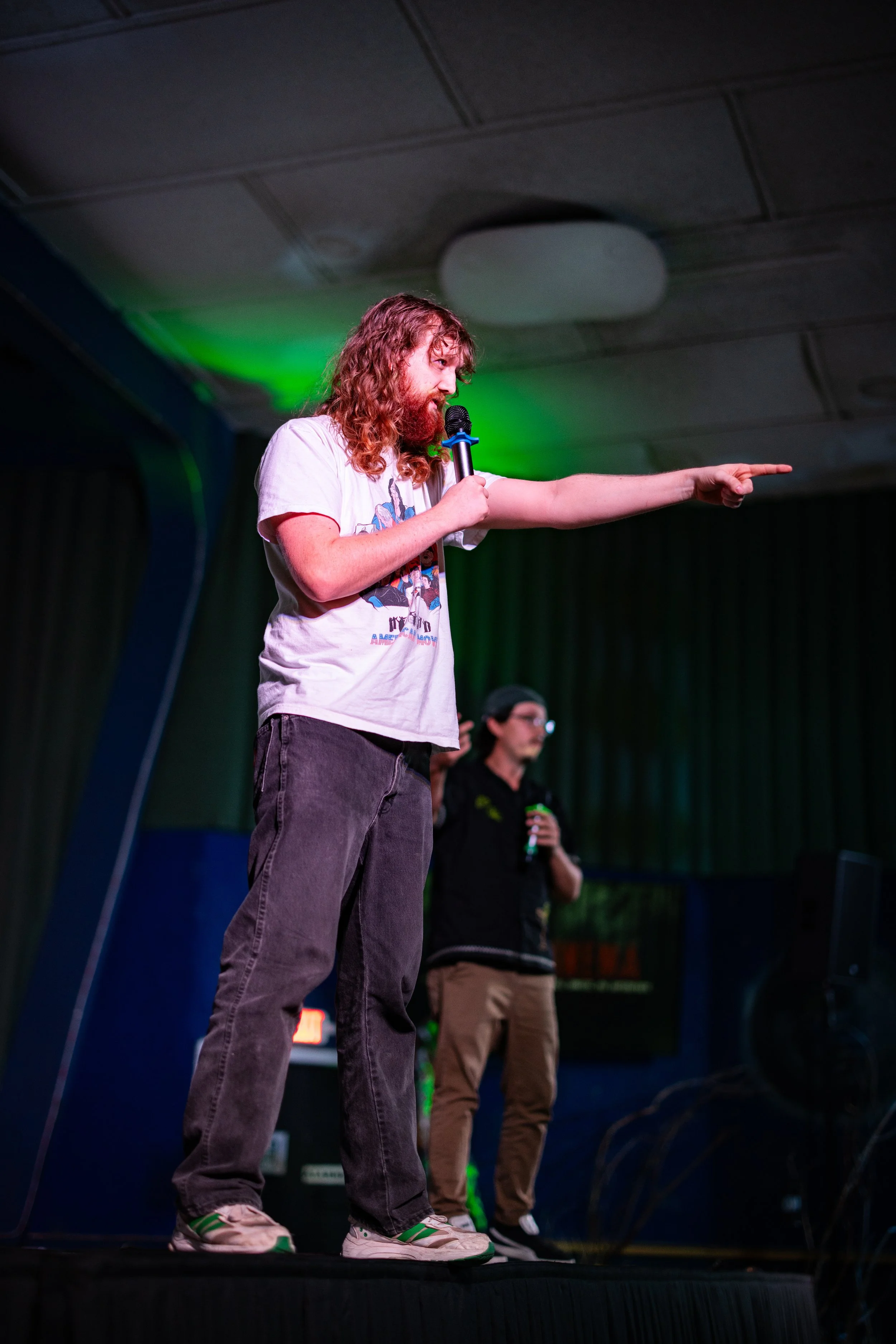 A man with long, curly hair and a beard is on stage holding a microphone in his right hand and pointing to the right with his left hand. He is wearing a white graphic T-shirt, dark jeans, and sneakers. There is a woman in the background holding a bev