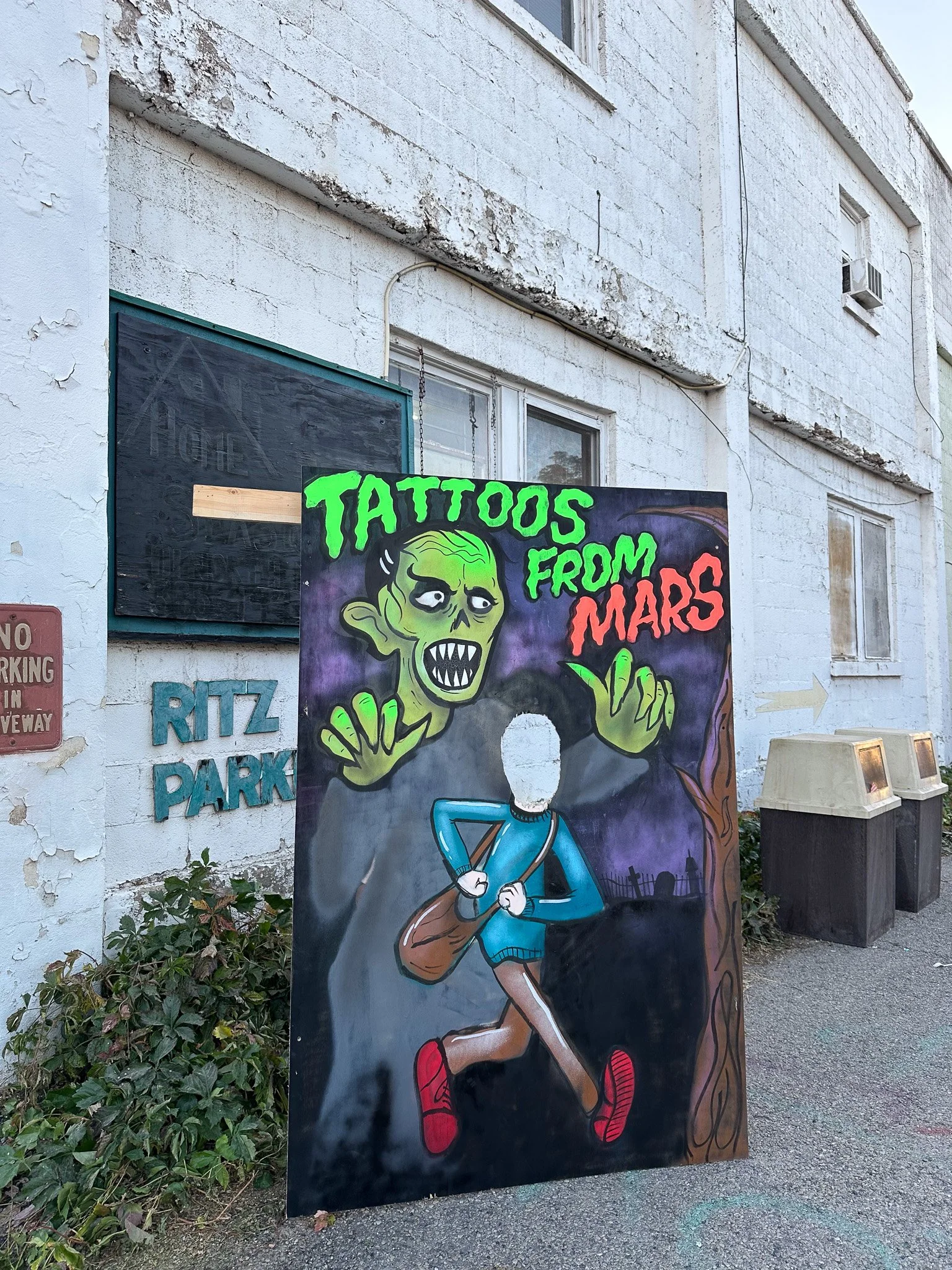 A colorful outdoor advertisement board for 'Tattoos from Mars,' featuring a cartoon green alien with sharp teeth and big eyes, reaching out over a person walking with a backpack, set against a dark purple background with a spooky, alien landscape. Th