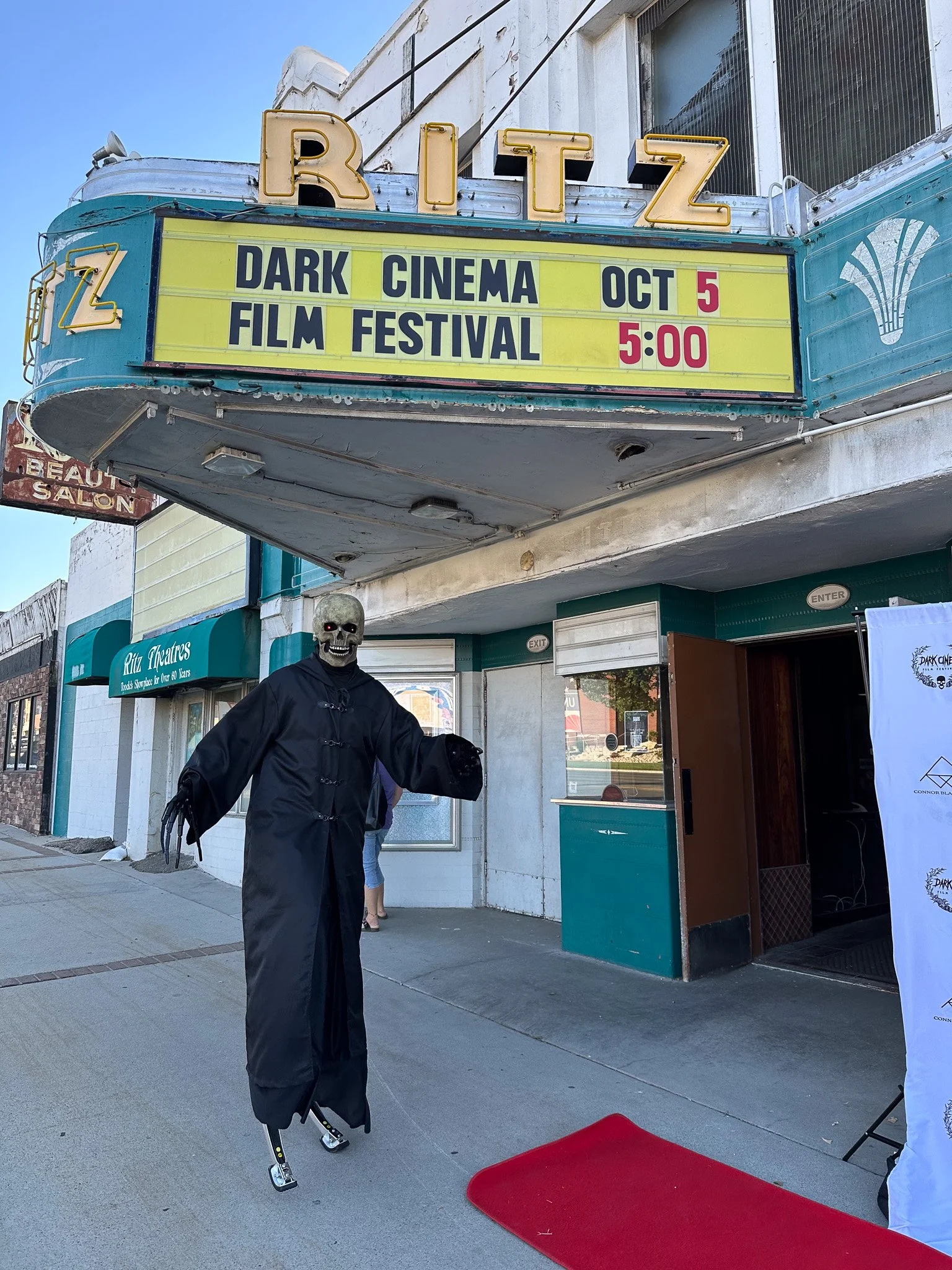 Skelton figure on stilts dressed in black coat with a skeleton mask standing outside a theater marquee advertising the Dark Cinema Film Festival on October 5 at 5:00 PM.