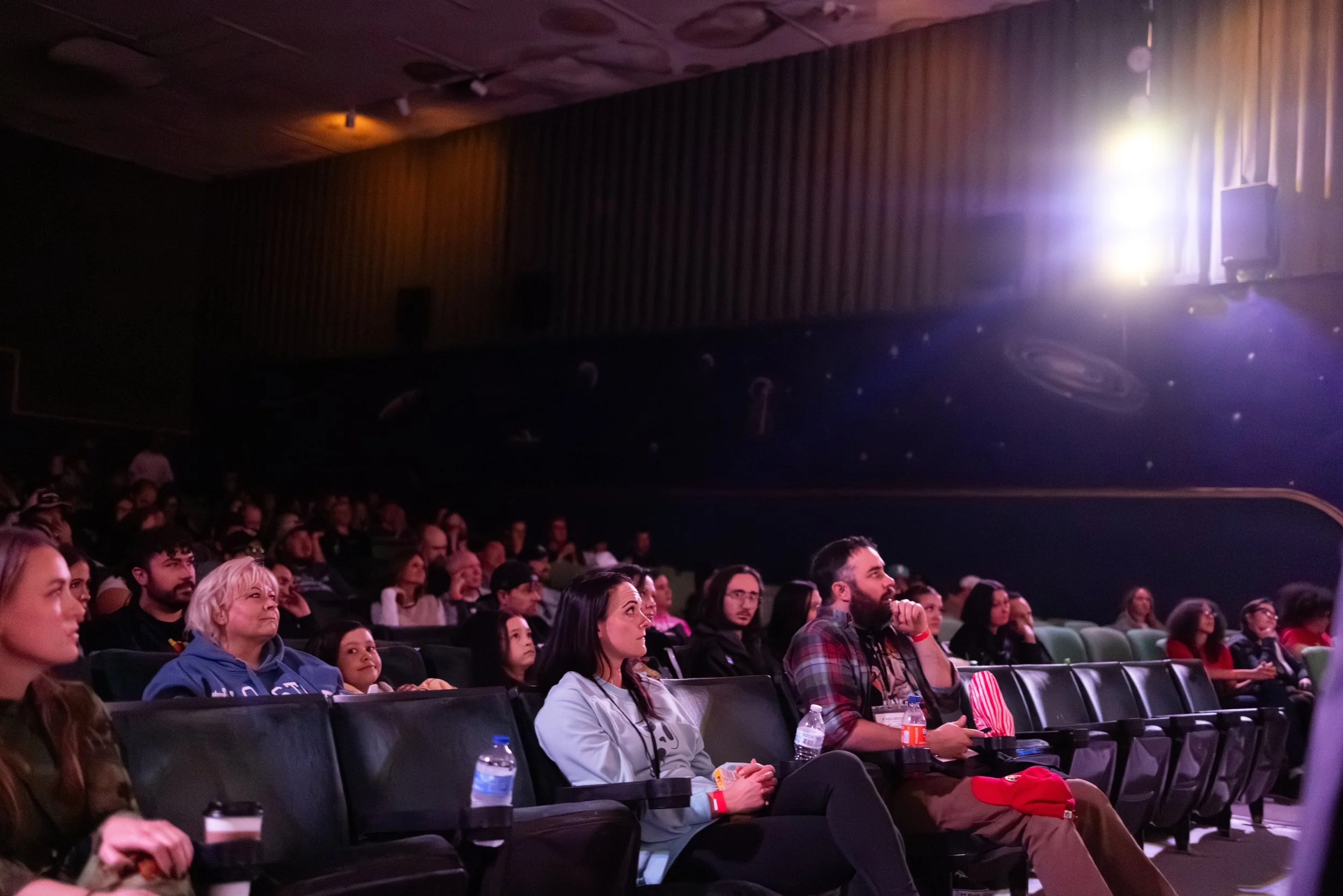 Audience seated in a dark theater watching a presentation or film, some with water bottles and popcorn, illuminated by dim stage lighting.