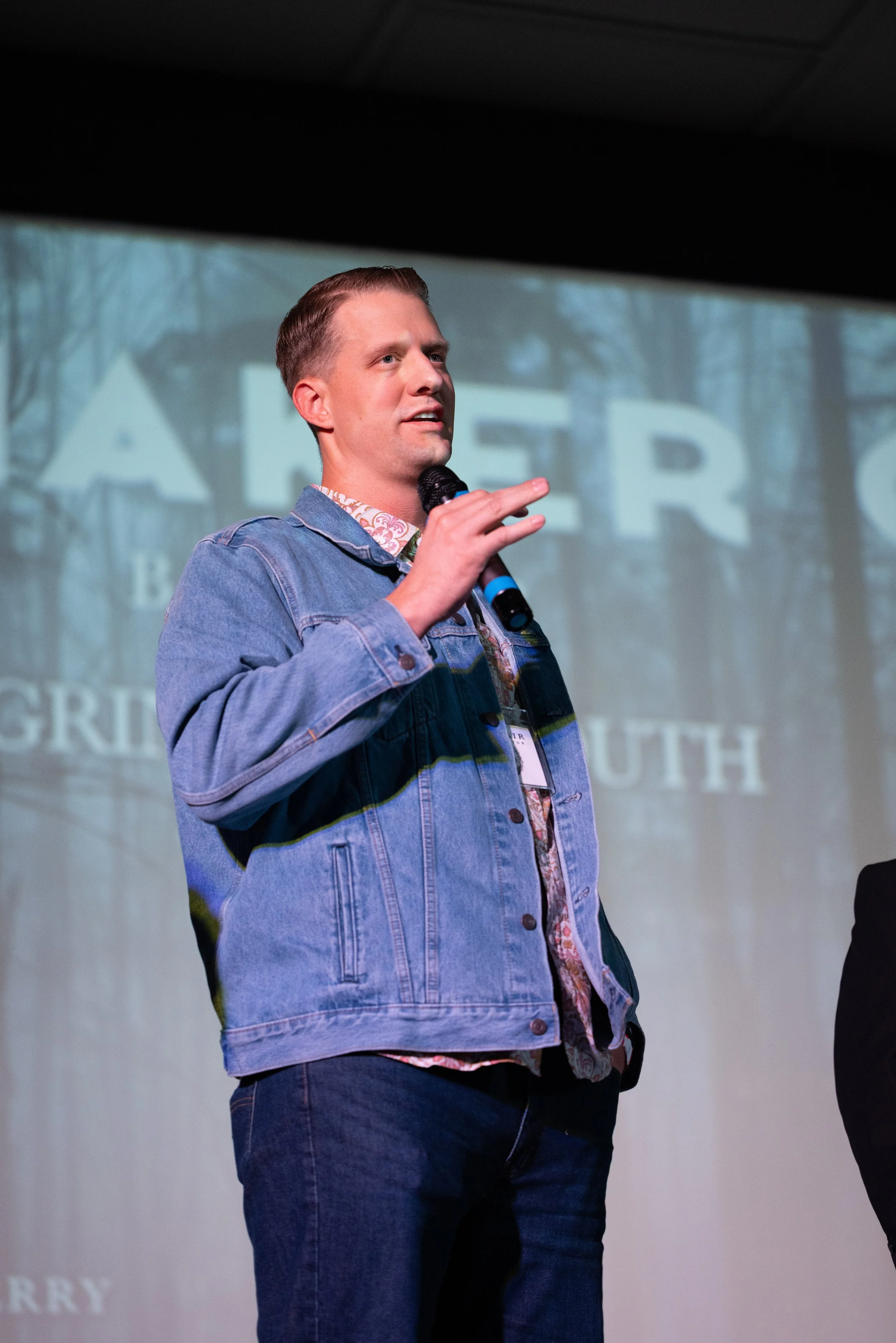 A young man with short hair, wearing a denim jacket and patterned shirt, holding a microphone and speaking on stage with a background featuring the word 'AFTER' and the words 'GRO', 'UTH', indicating possibly a presentation or event.