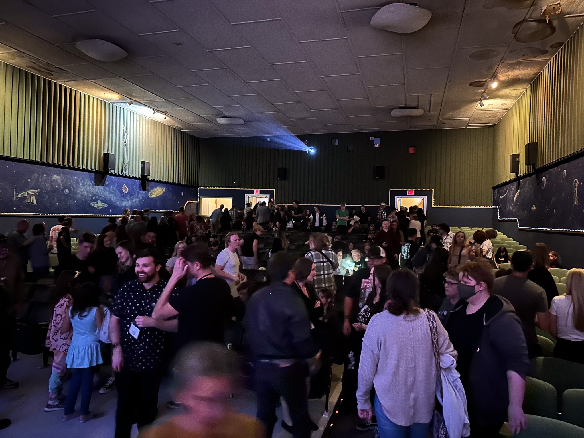 Crowd of people inside a movie theater or auditorium, with some standing, talking, and seated. The walls are decorated with space-themed murals.