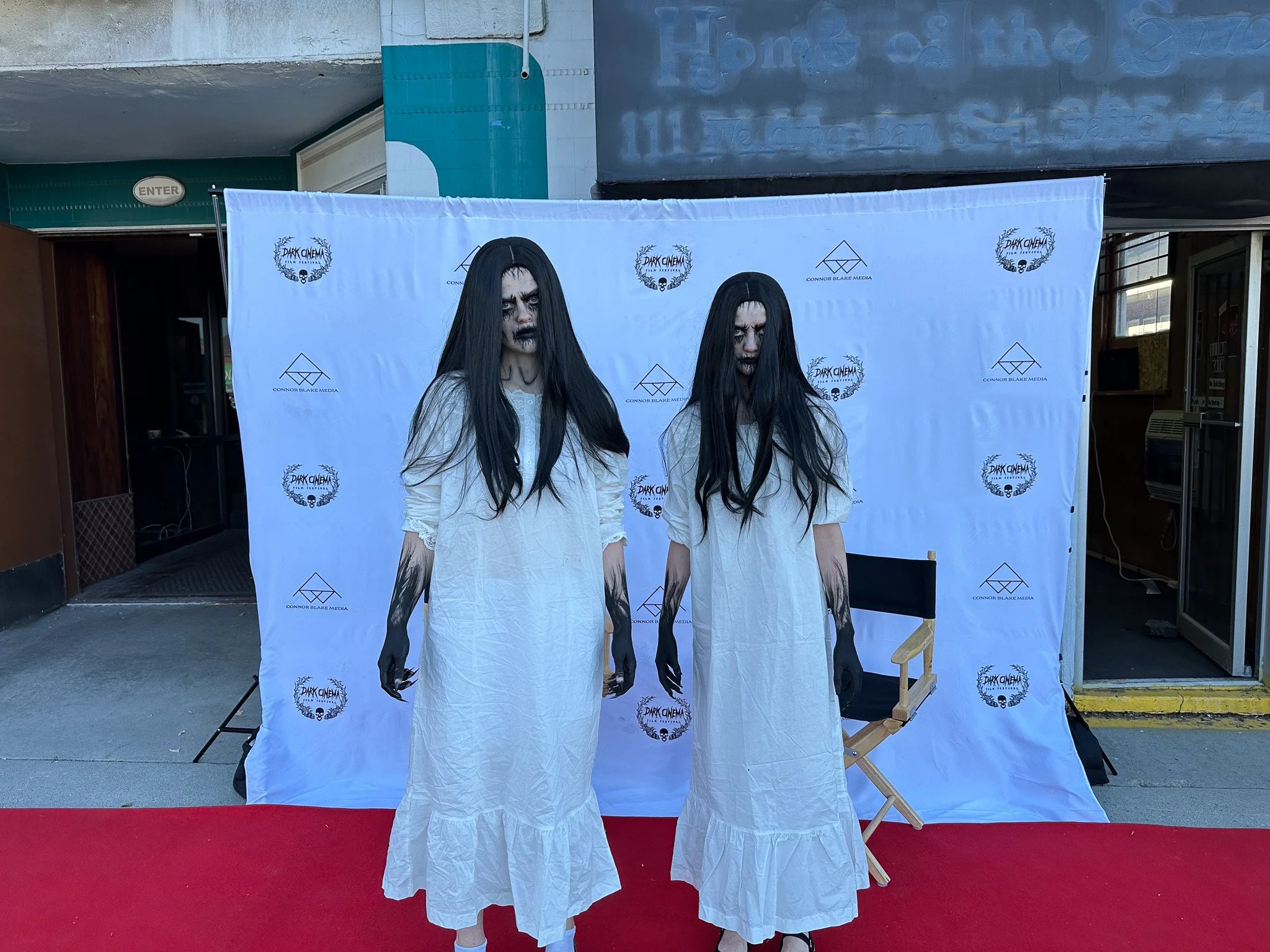 Two women dressed as horror movie ghosts with long black hair, pale face makeup, black lips, and white nightgown costumes, standing in front of a step-and-repeat backdrop with event logos at an outdoor venue.