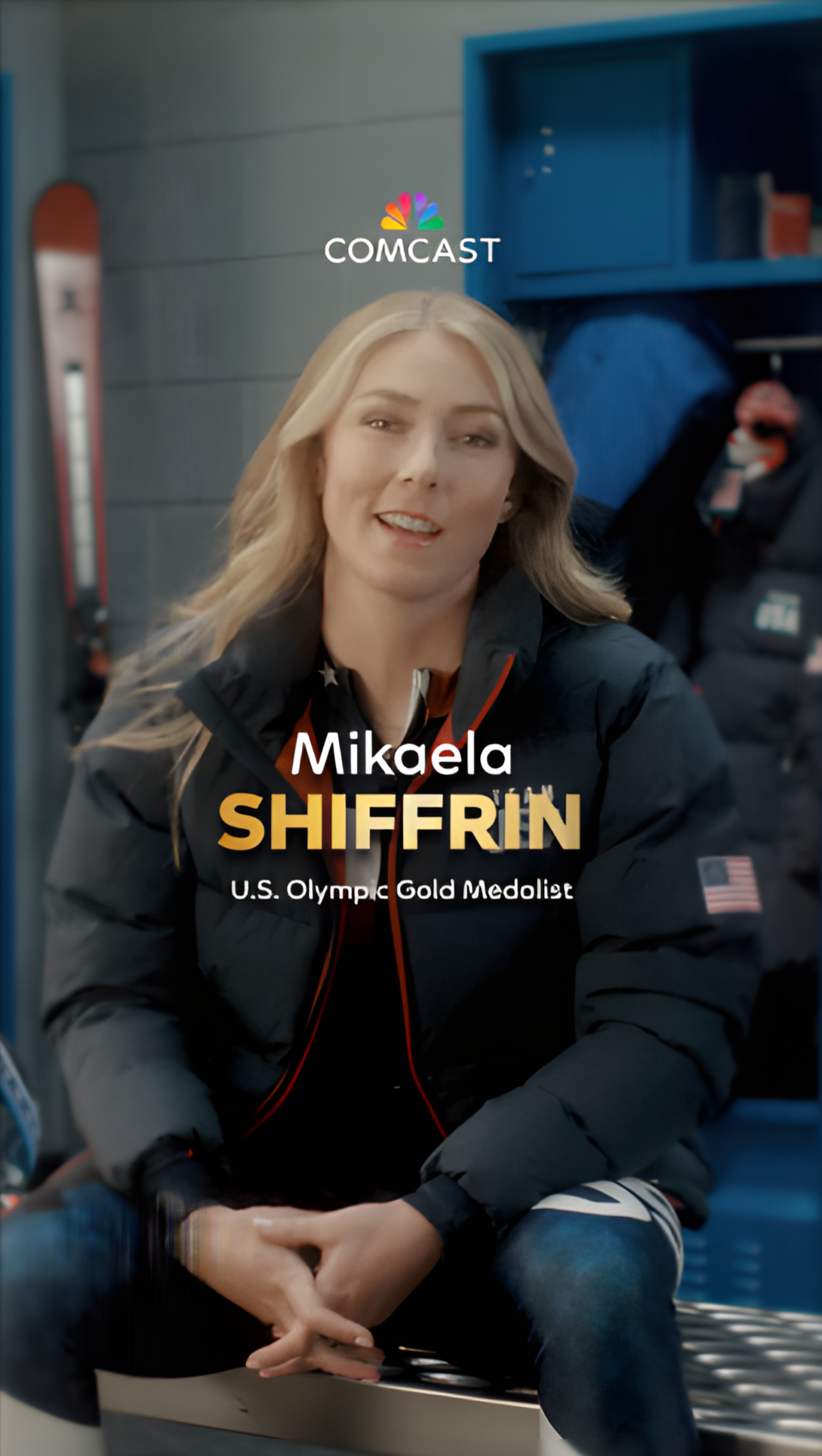 A woman with long blonde hair, sitting and smiling in a sportswear jacket with an American flag patch, in an indoor setting with lockers and shelves in the background.