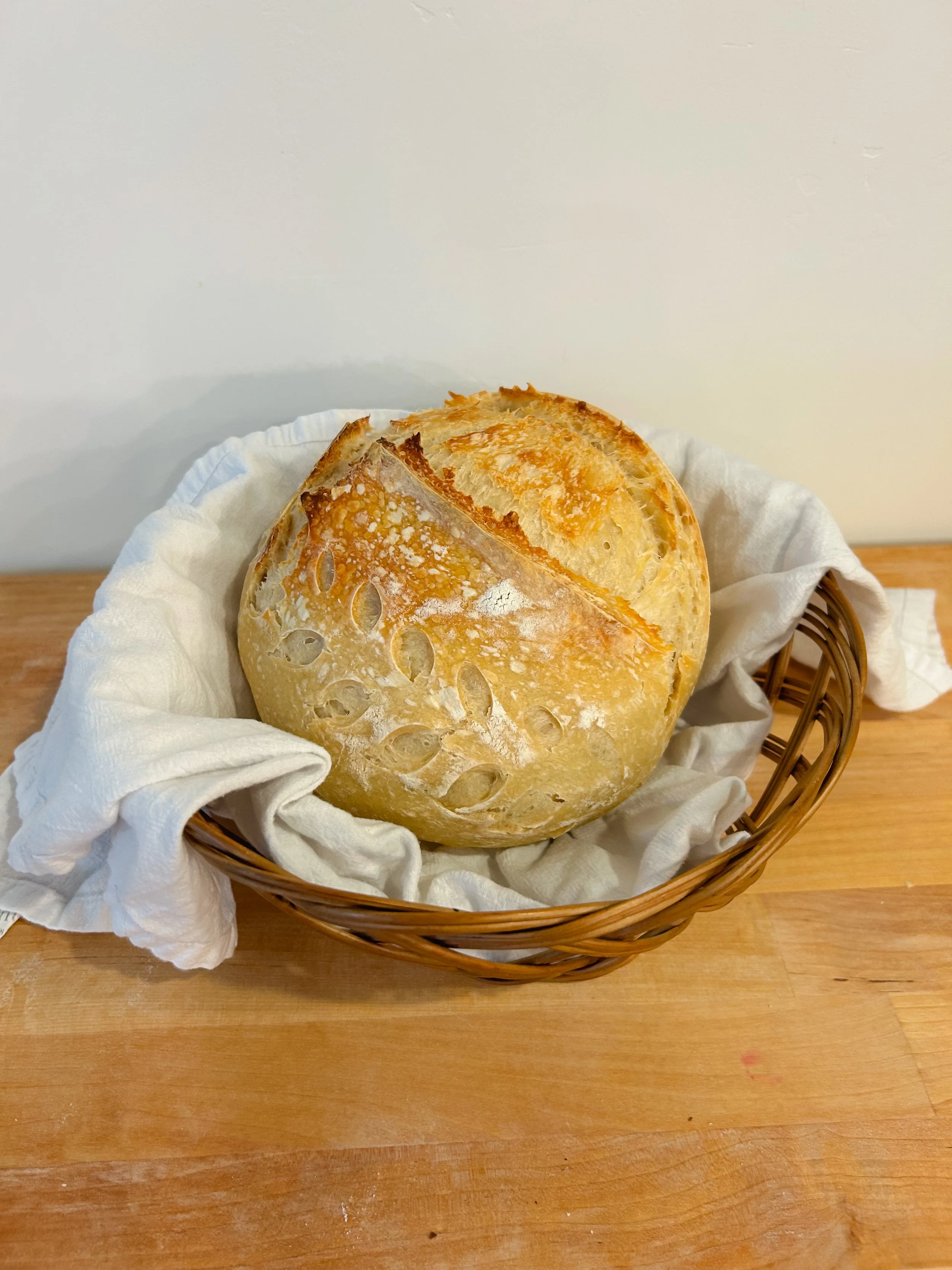 Round loaf of freshly baked asiago bread in a wicker basket lined with a white cloth on a wooden surface.