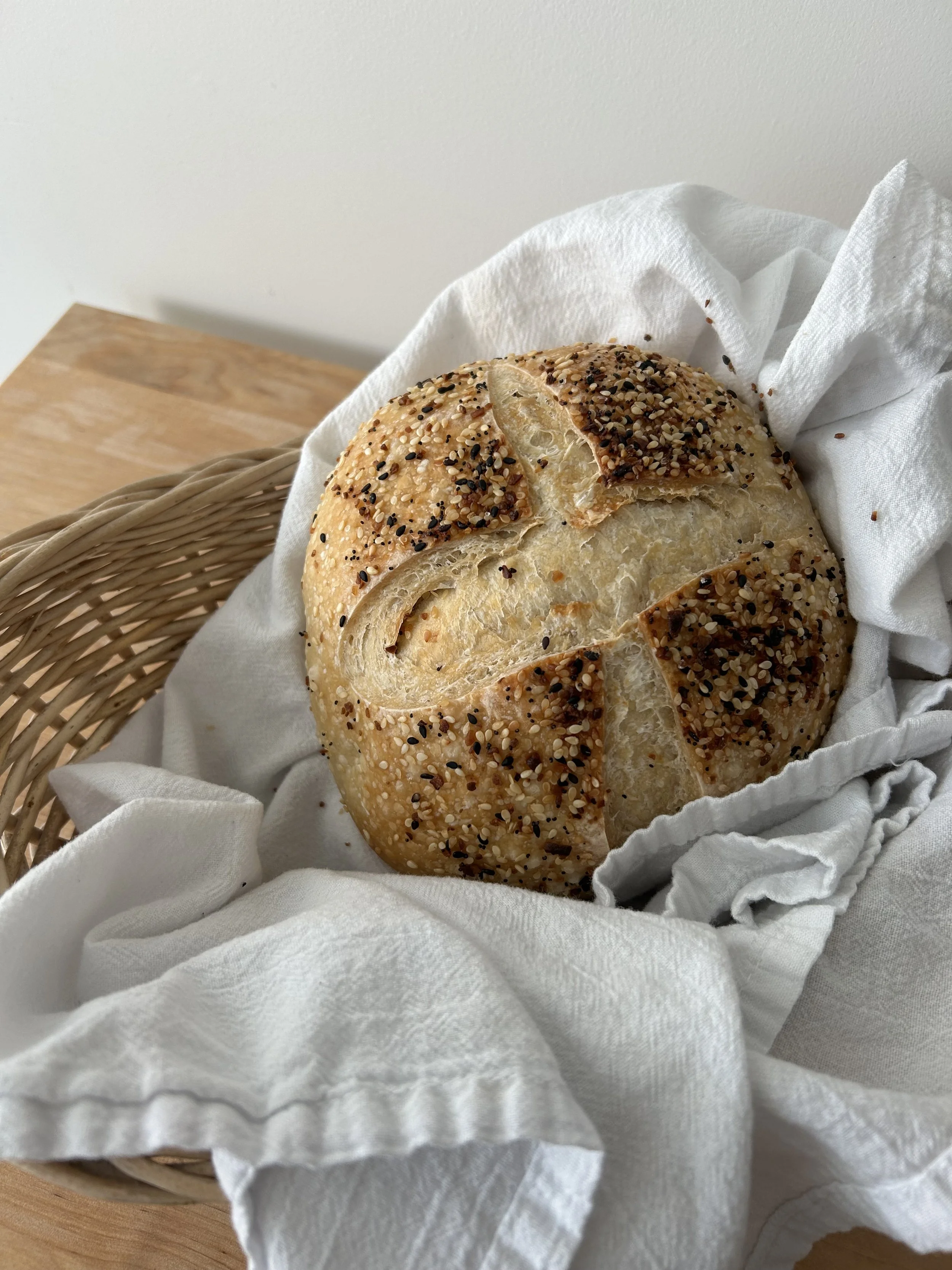 Round loaf of bread topped with seeds in a wicker basket lined with white cloth.