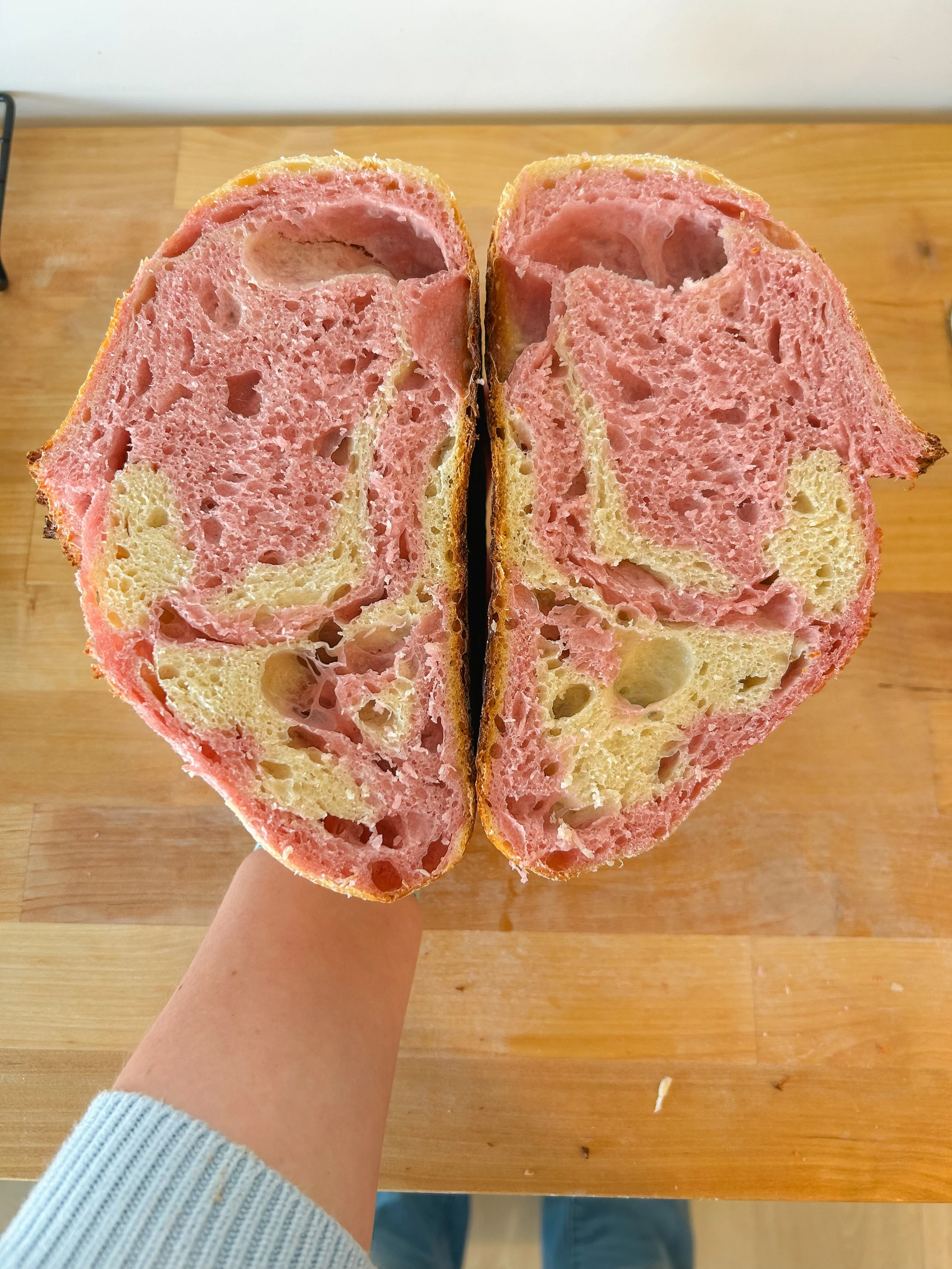 Close-up of a halved pink and yellow bagel with a dense, airy interior, held over a wooden table.