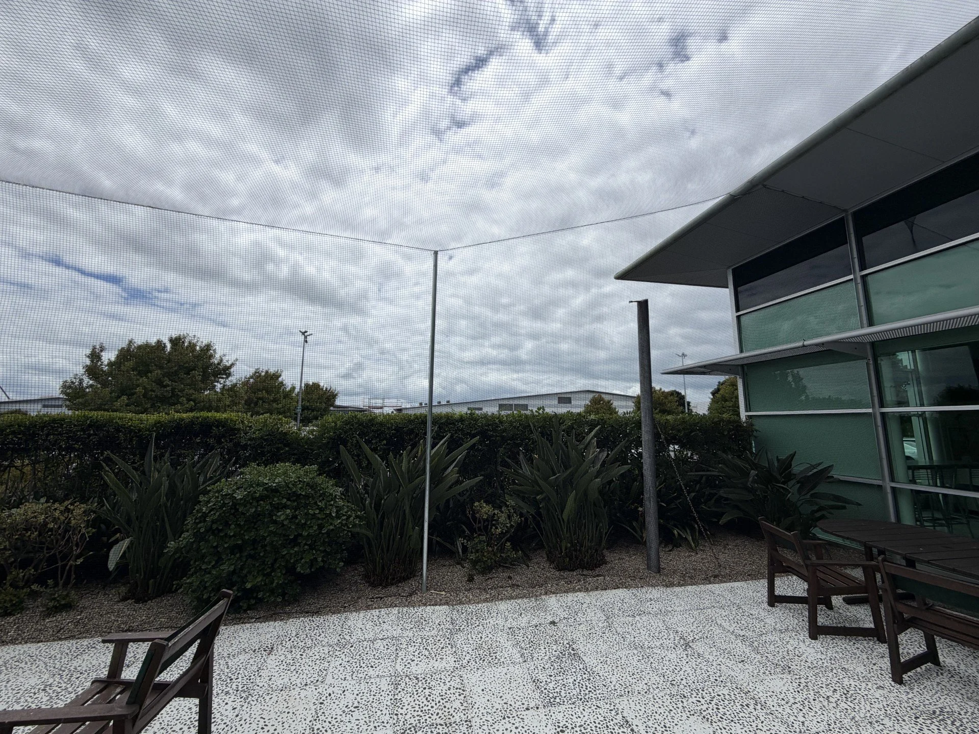 Outdoor patio area with wooden chairs, green bushes, a building with glass windows, and a net above, under cloudy sky.