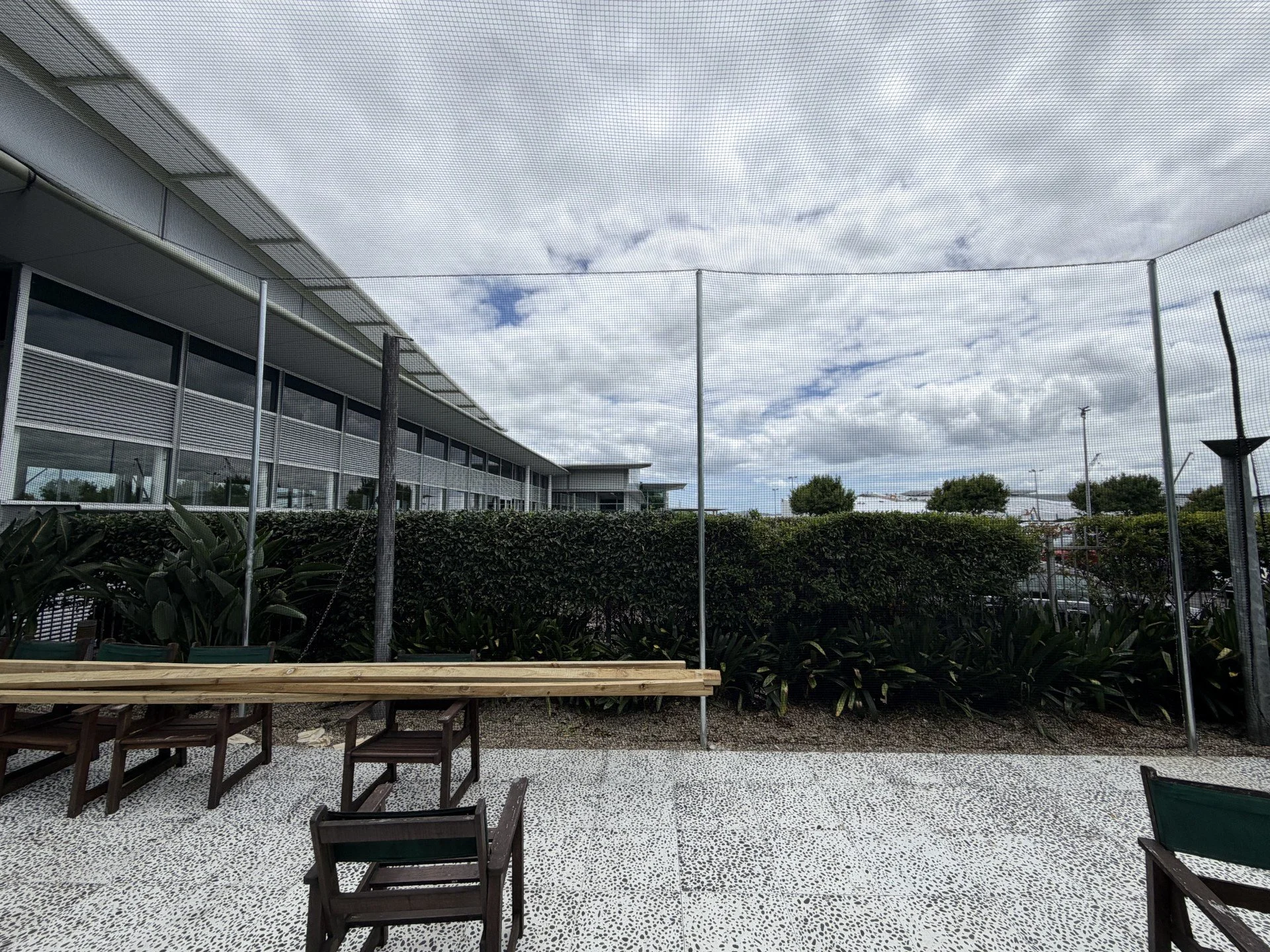 Outdoor patio area with wooden tables and chairs, tall hedge, mesh netting overhead, cloudy sky, modern building in background with glass windows.
