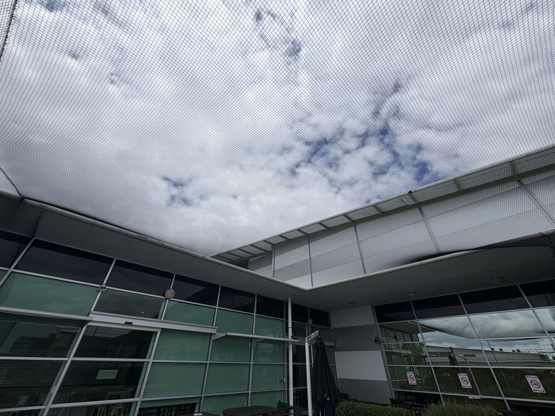 Overcast sky seen through a protective mesh roof over a modern commercial building with glass windows and an outdoor seating area. Showing bird netting to stop birds from getting inside building.