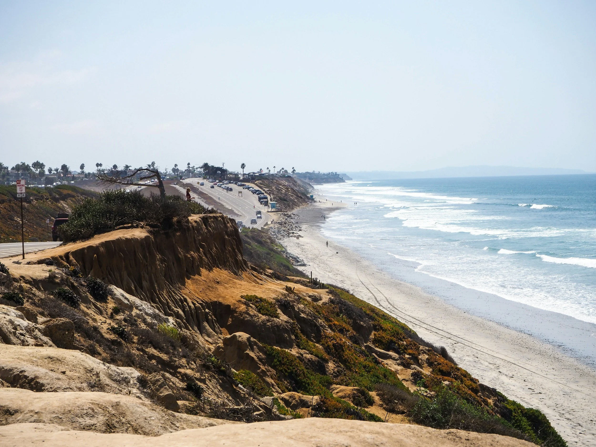 View of a coastal road running along cliffs with parked cars, a sandy beach, and the ocean waves on a clear day.