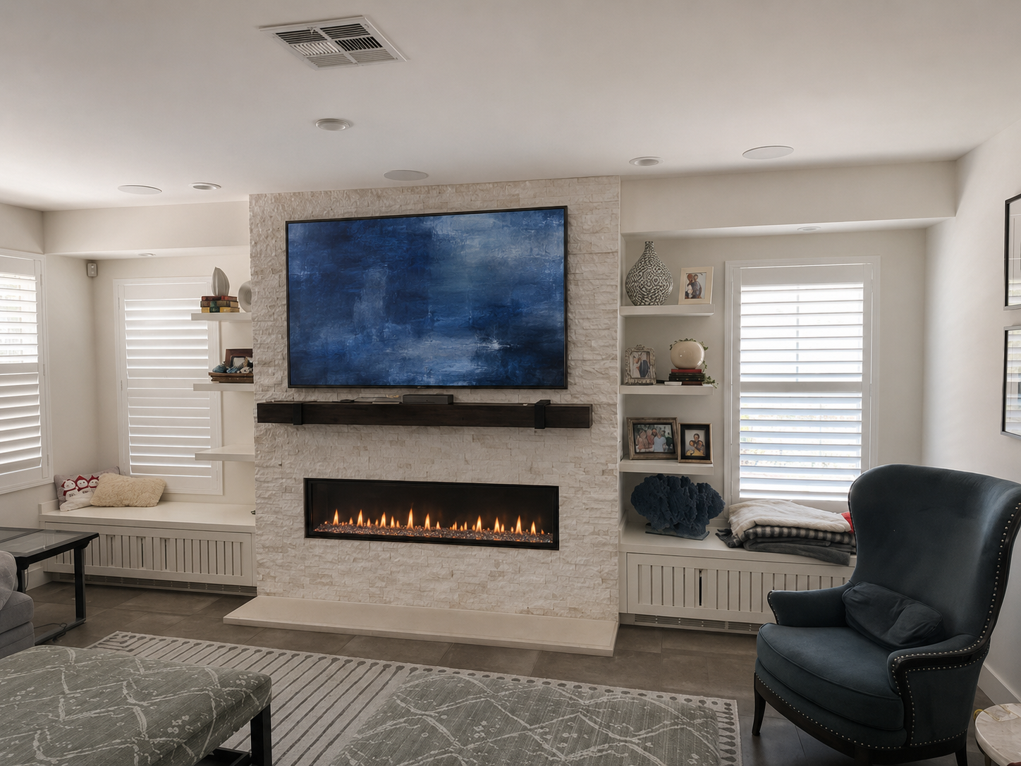Living room with a white brick fireplace, a large flat-screen TV above it, a cozy navy armchair, glass coffee table, built-in shelves with decorative items, and windows with white plantation shutters.