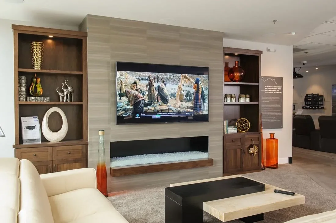 This is a Great Room system in Carlsbad featuring a porcelain clad tile bump out for a electric linear fireplace with flanking walnut cabinets.  Flush mount speakers with the right front grill removed so you can see it.  Equipment hidden in convectio