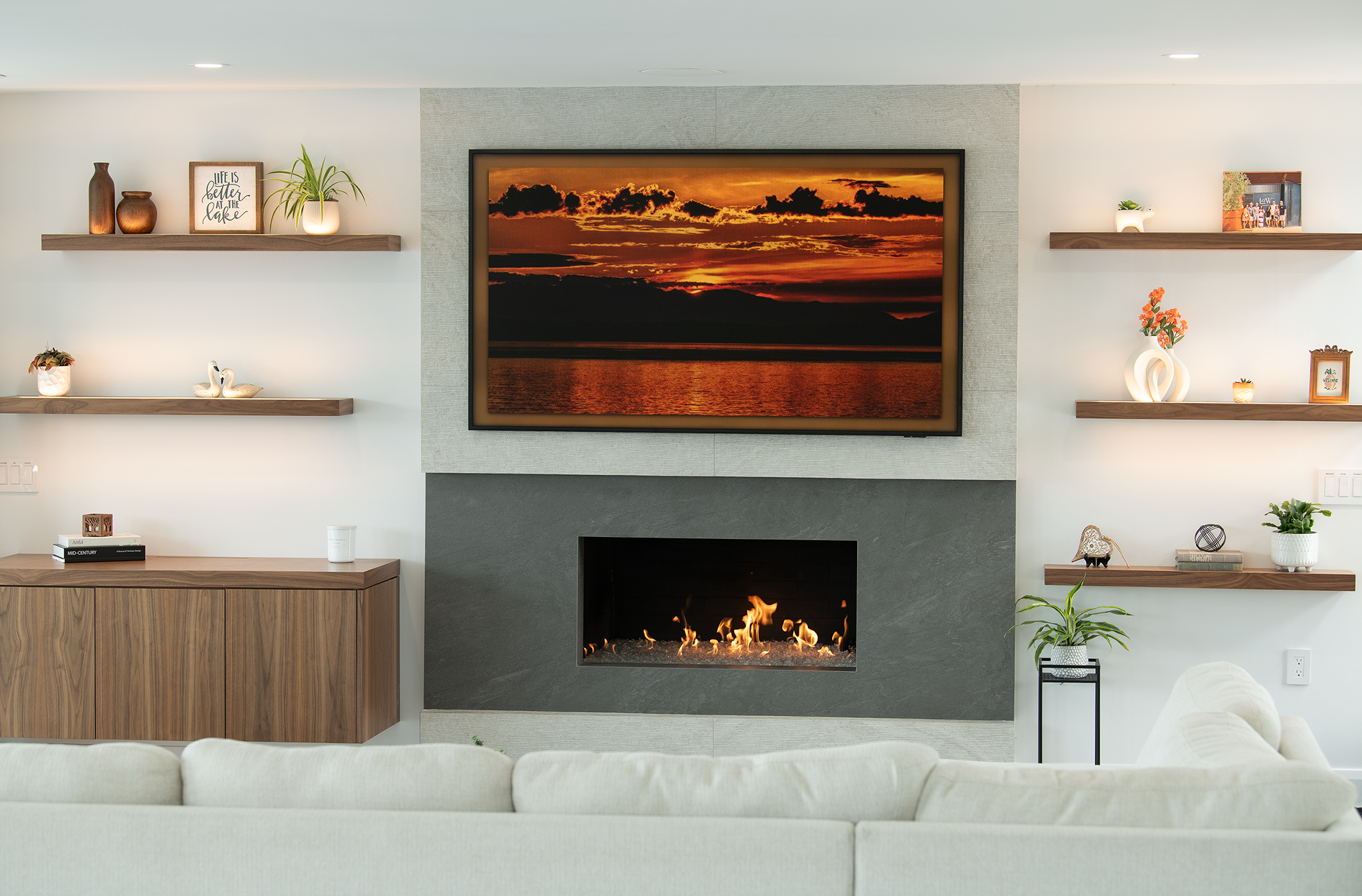 A modern living room with a fireplace and a large framed sunset picture above it, surrounded by wall-mounted shelves with decorative items like vases, plants, and photos.