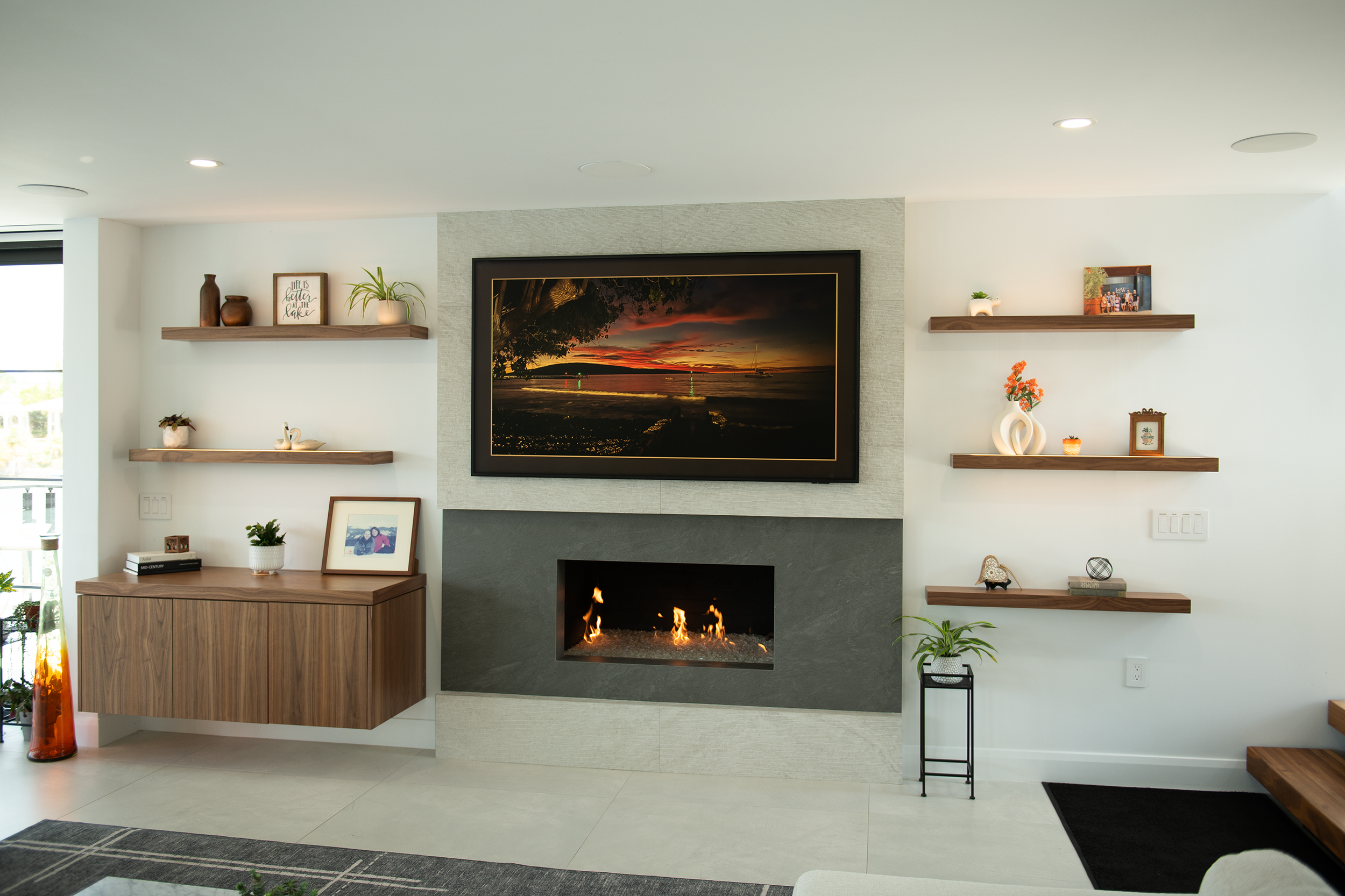 Living room with a wall-mounted TV displaying sunset over a lake, a fireplace under the TV, and decorated shelves with plants, framed photos, and vases.