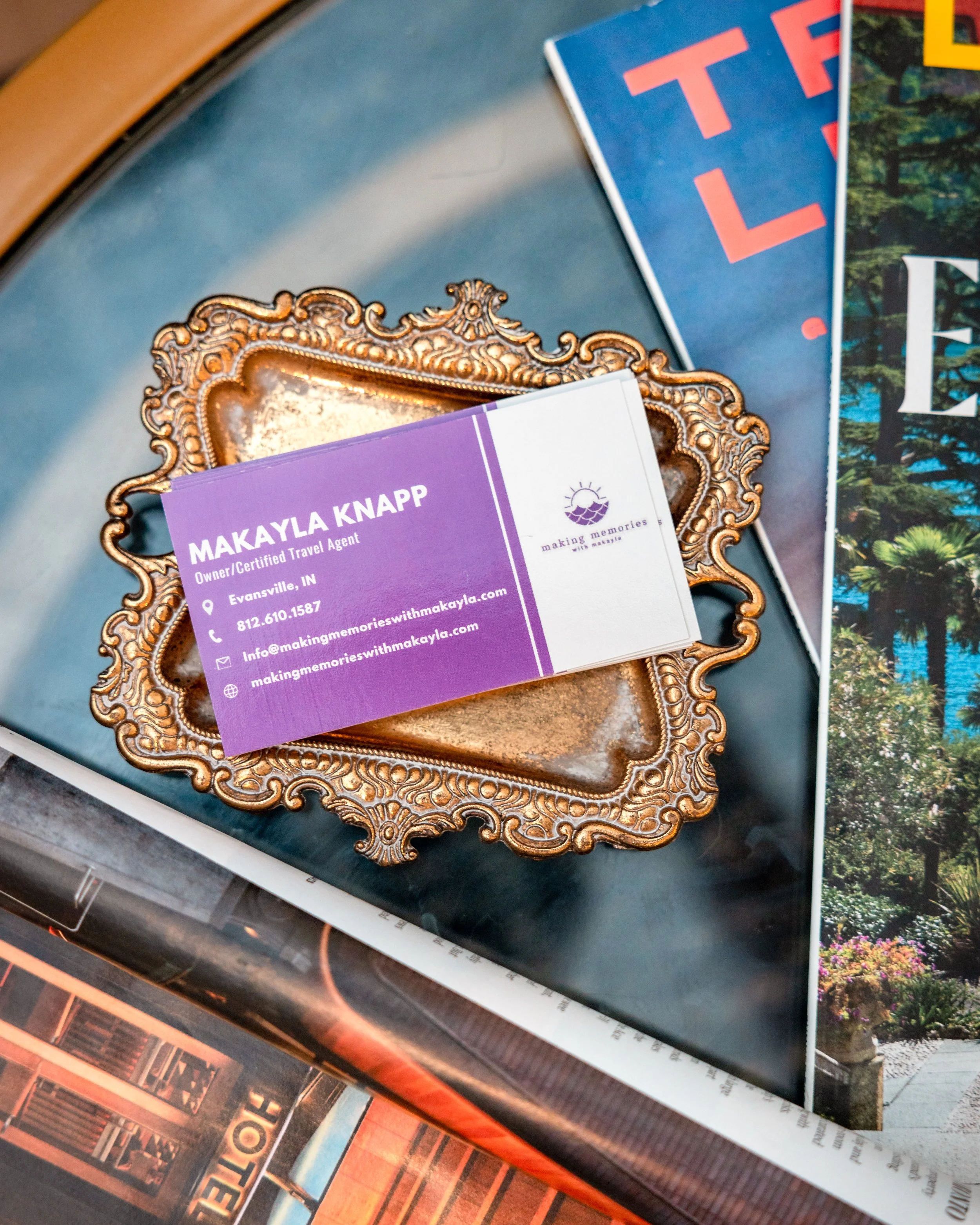 A business card for Makayla Knapp, a certified travel agent from Evansville, Indiana, resting on an ornate gold tray on a blue table surrounded by magazines.