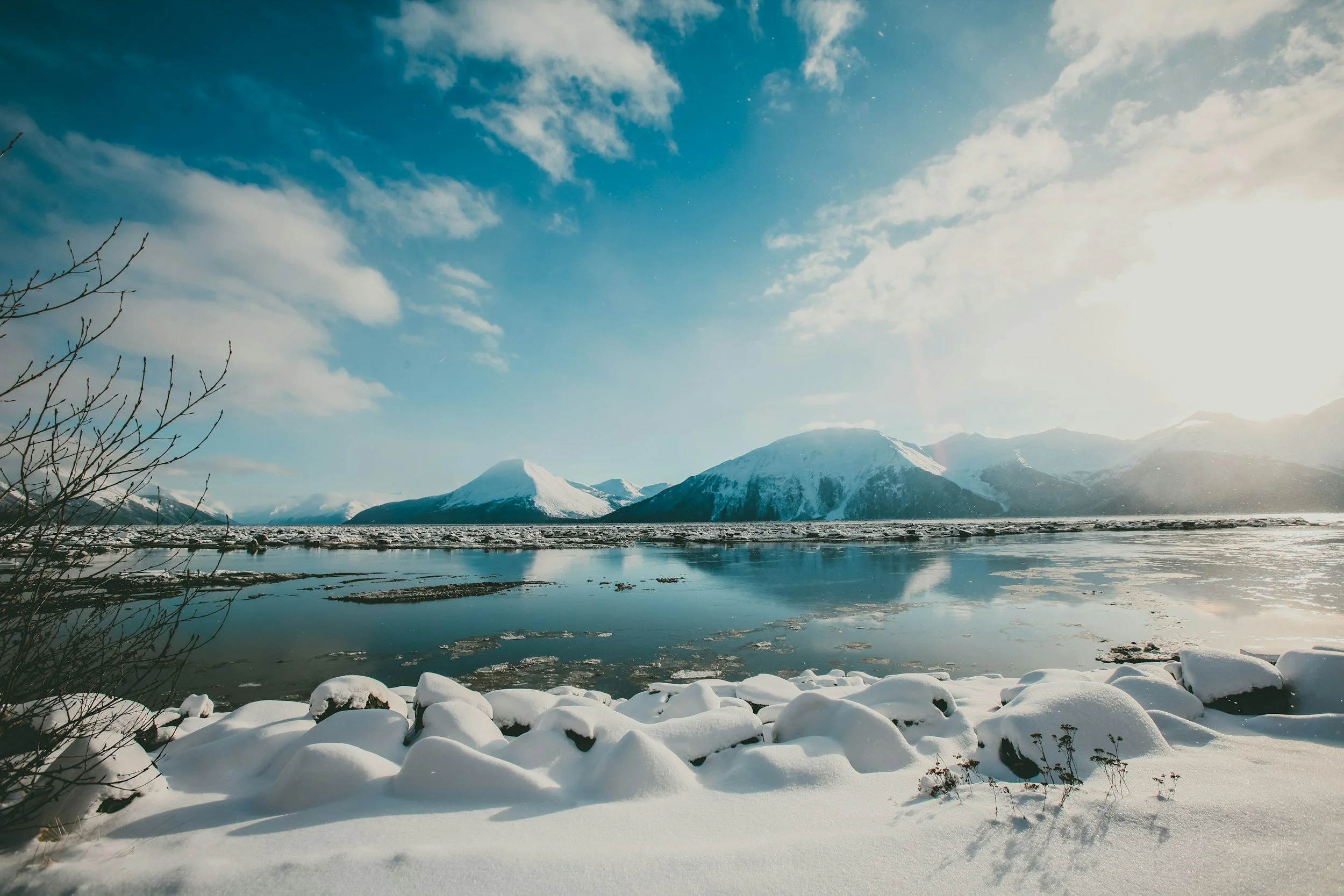 Snow-covered rocks and shoreline in the foreground, a partially frozen lake reflecting snow-capped mountains in the background, under a partly cloudy sky with sunlight.