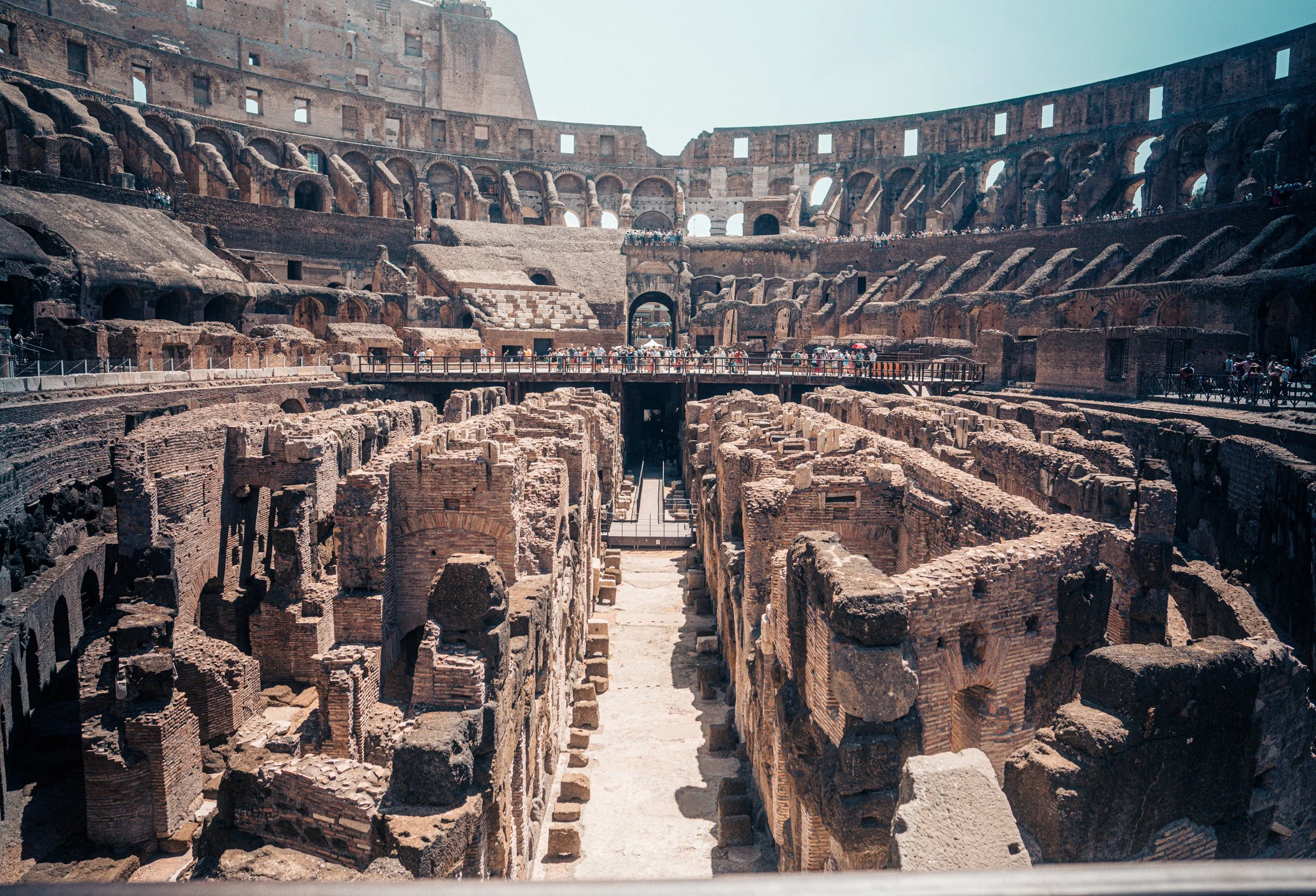 The interior of the ancient Roman Colosseum with exposed brick and stone ruins, multiple levels of arches, and a large crowd of tourists exploring the archaeological site.